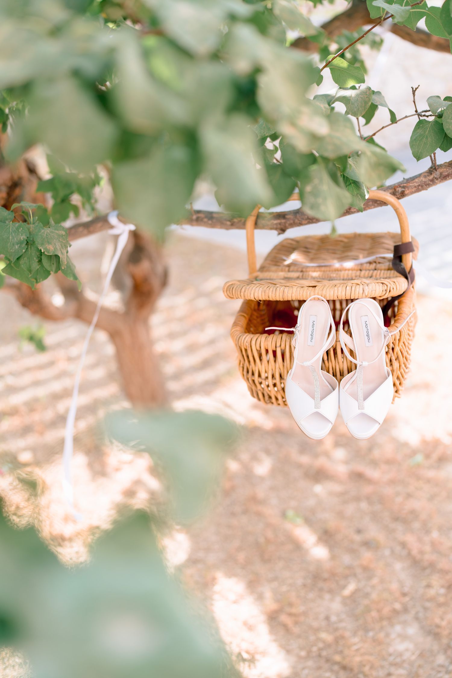 Delicate white bridal sandals hanging from a wicker basket among green leaves.
