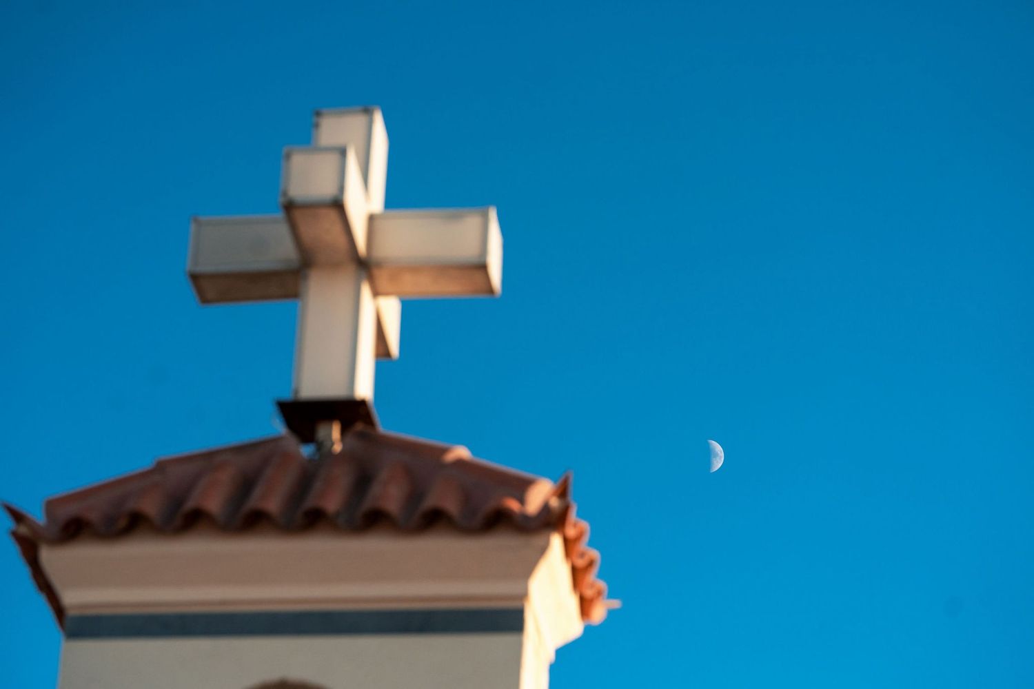 White cross atop a church steeple against a bright blue sky with a crescent moon in view.