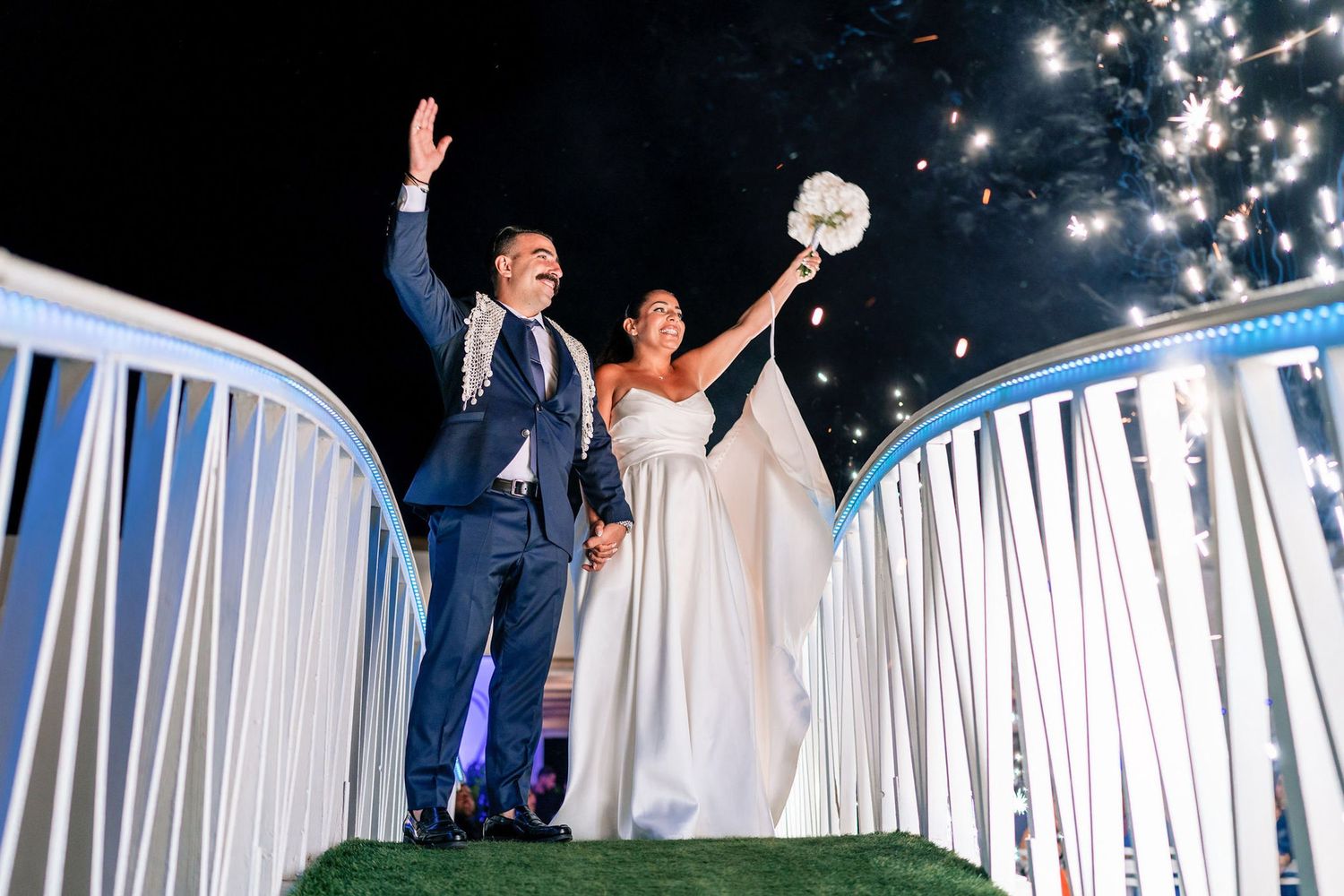 Wedding guests wave sparklers during an outdoor nighttime celebration on a white-railed deck.