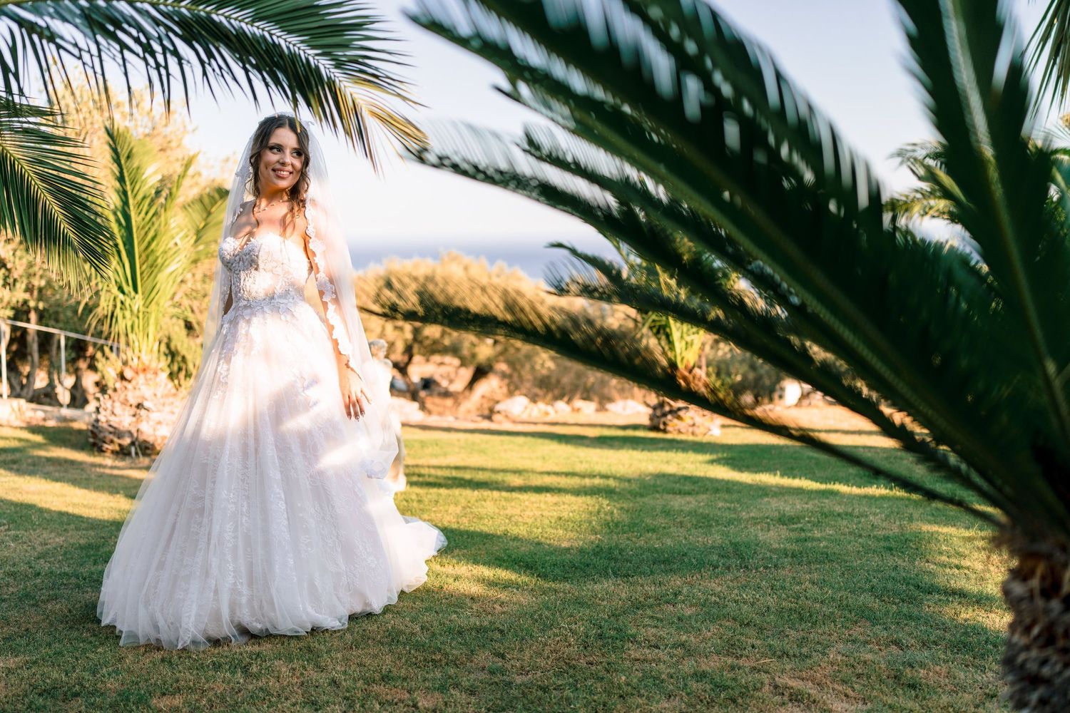 Bride in white ballgown poses between palm trees on manicured lawn.