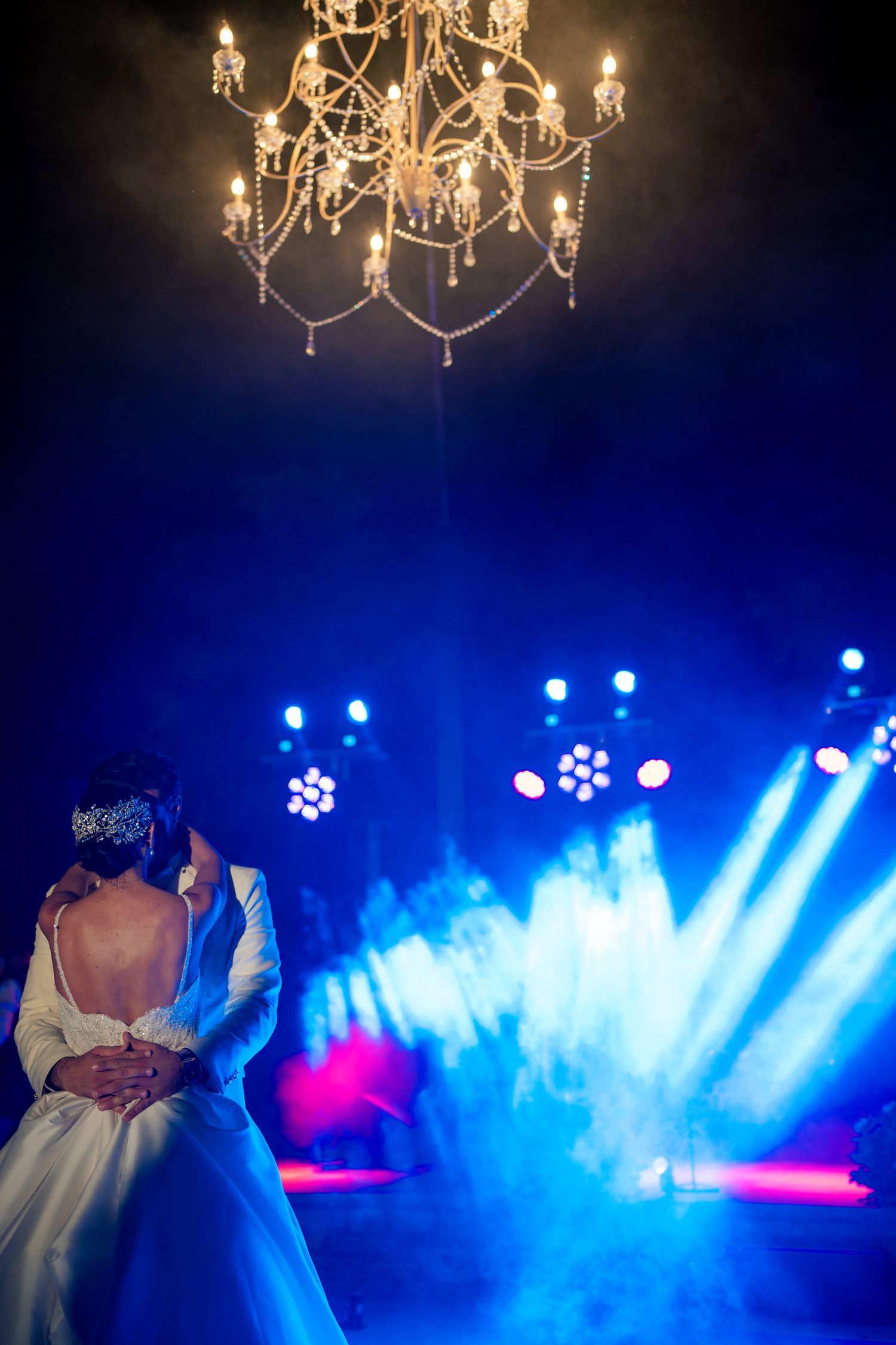 Wedding couple dances under ornate chandelier with dramatic blue stage lighting and fog effects.