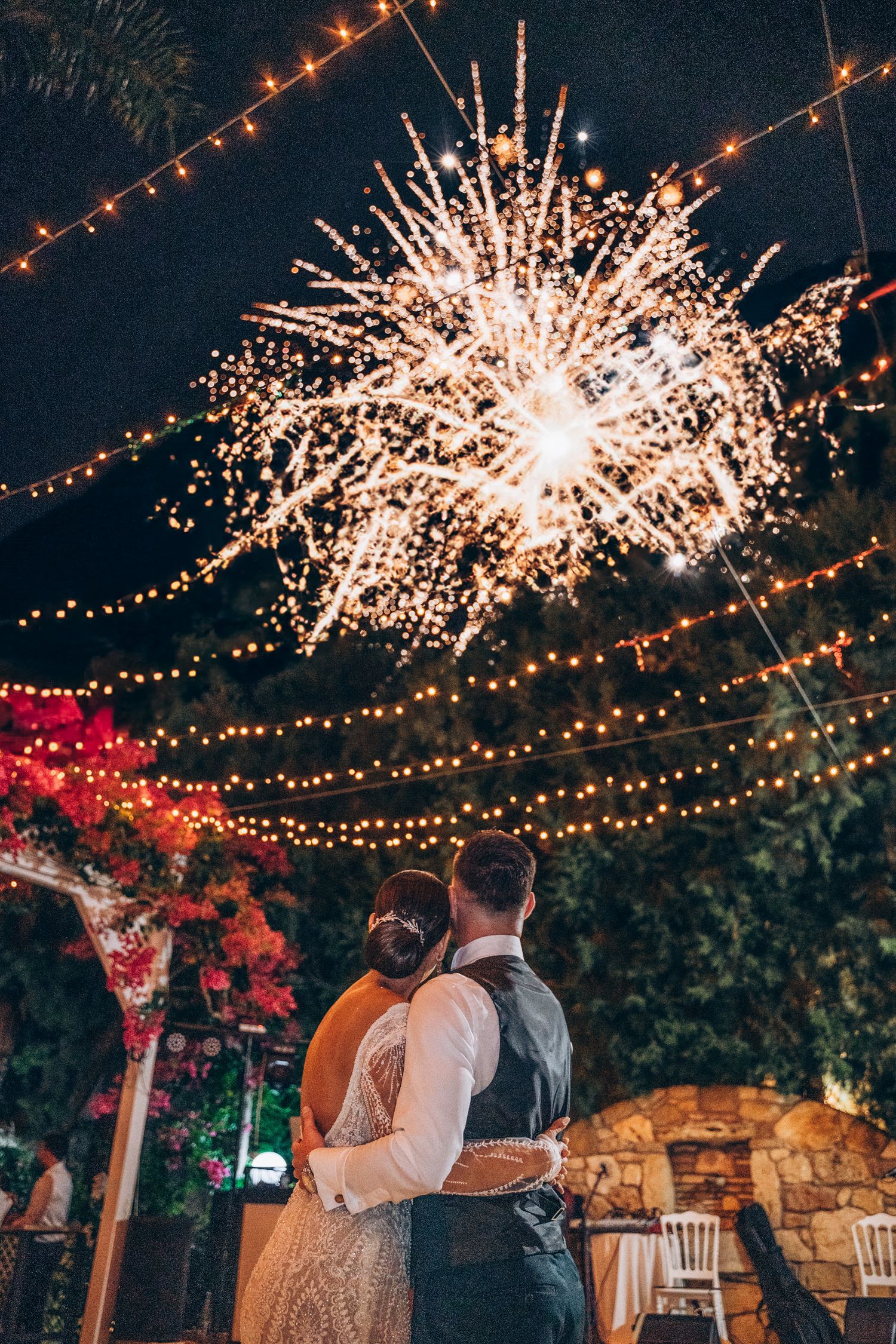 Outdoor wedding reception under twinkling string lights and fireworks creating a magical nighttime atmosphere.