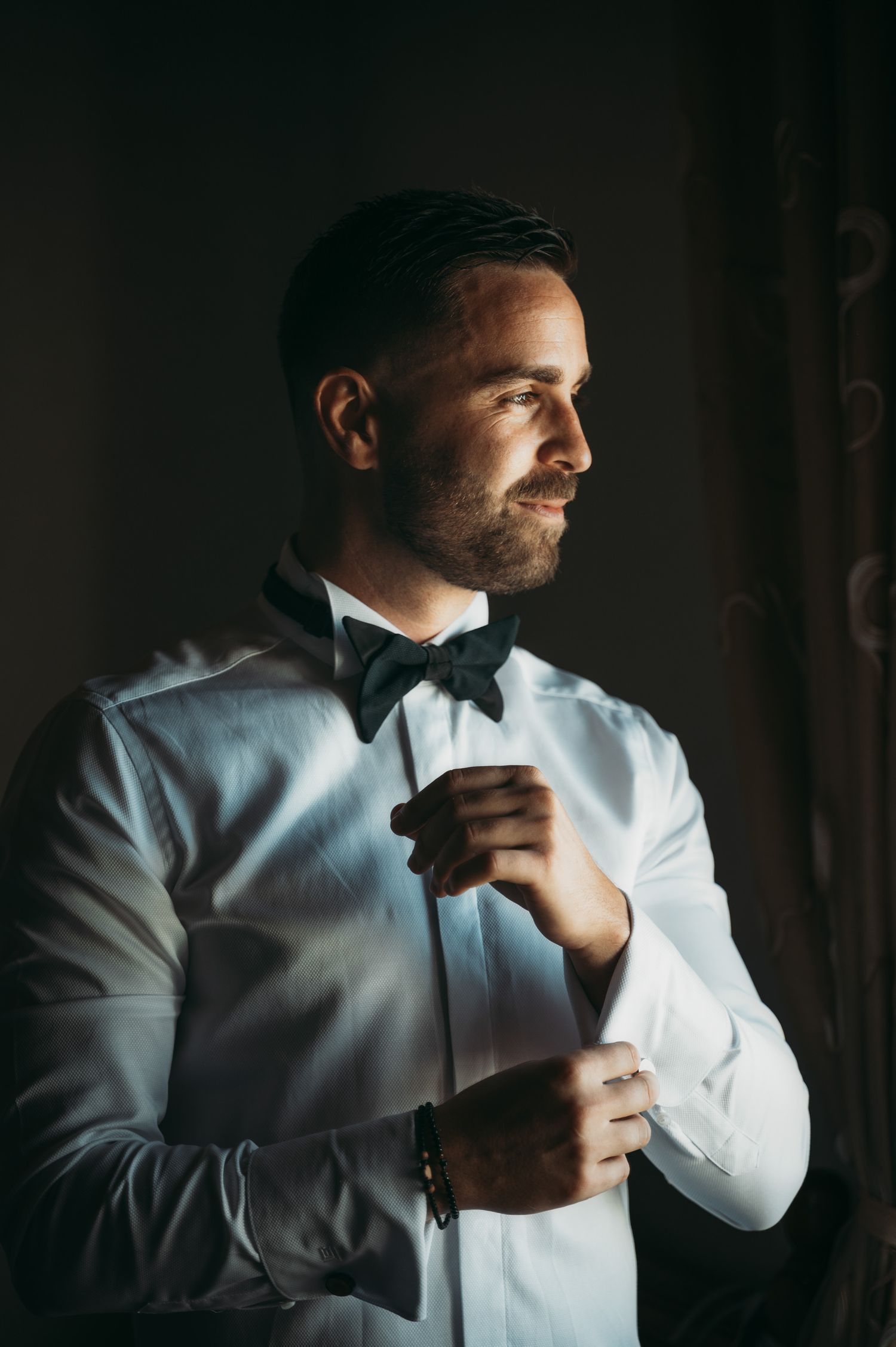 Groom adjusts tie and cuffs in dramatic window lighting.