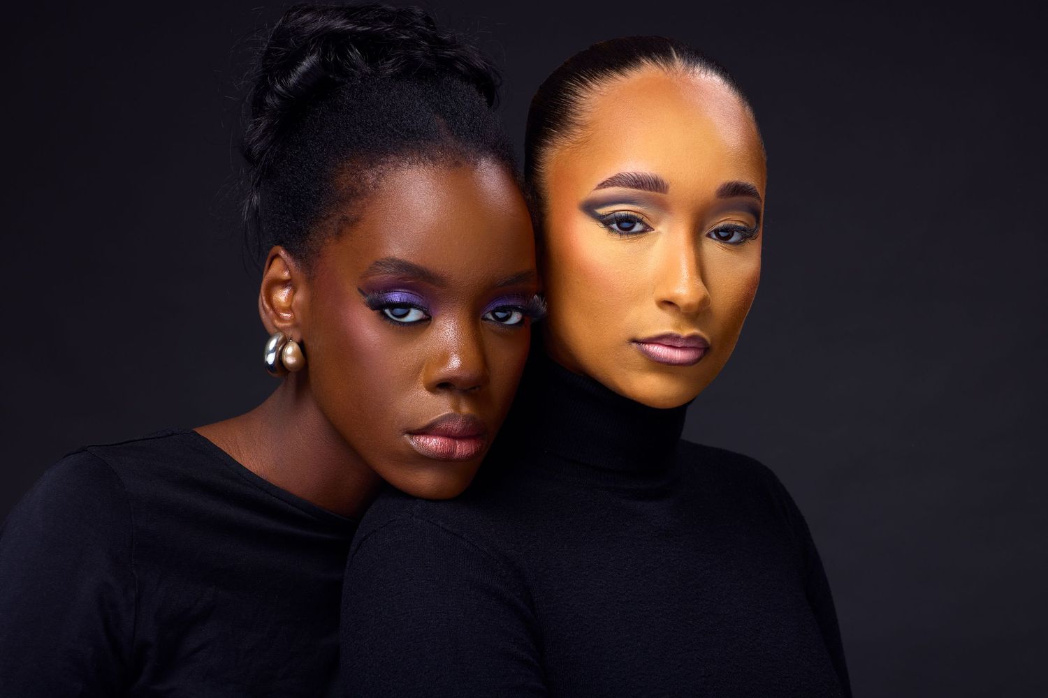Two Black women in black outfits with bold colorful makeup pose together against a dark background.