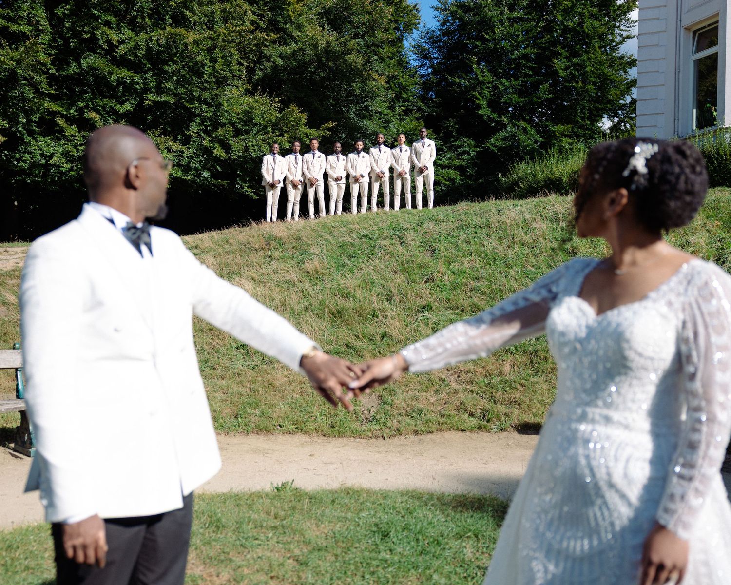 A wedding party dressed in white poses outdoors on a grassy slope during a sunny summer day.