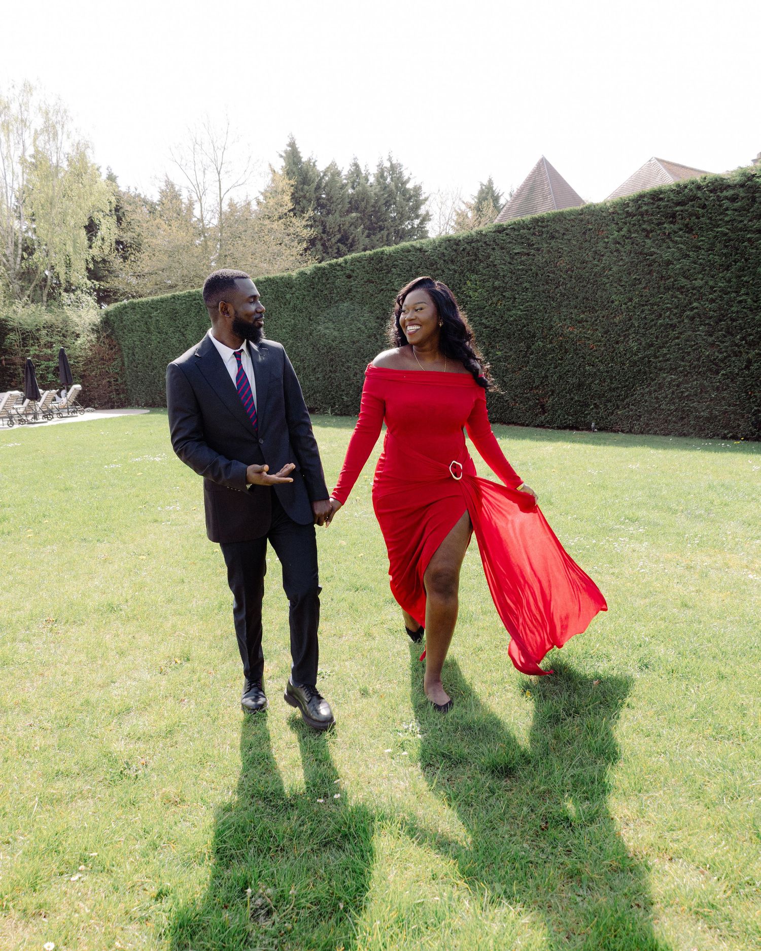 A couple walks hand-in-hand on green grass, man in dark suit, woman in flowing red off-shoulder dress, hedge backdrop.