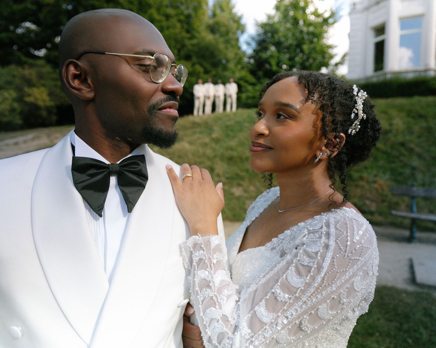 A romantic outdoor wedding portrait shows a couple in white formalwear embracing during their photoshoot.