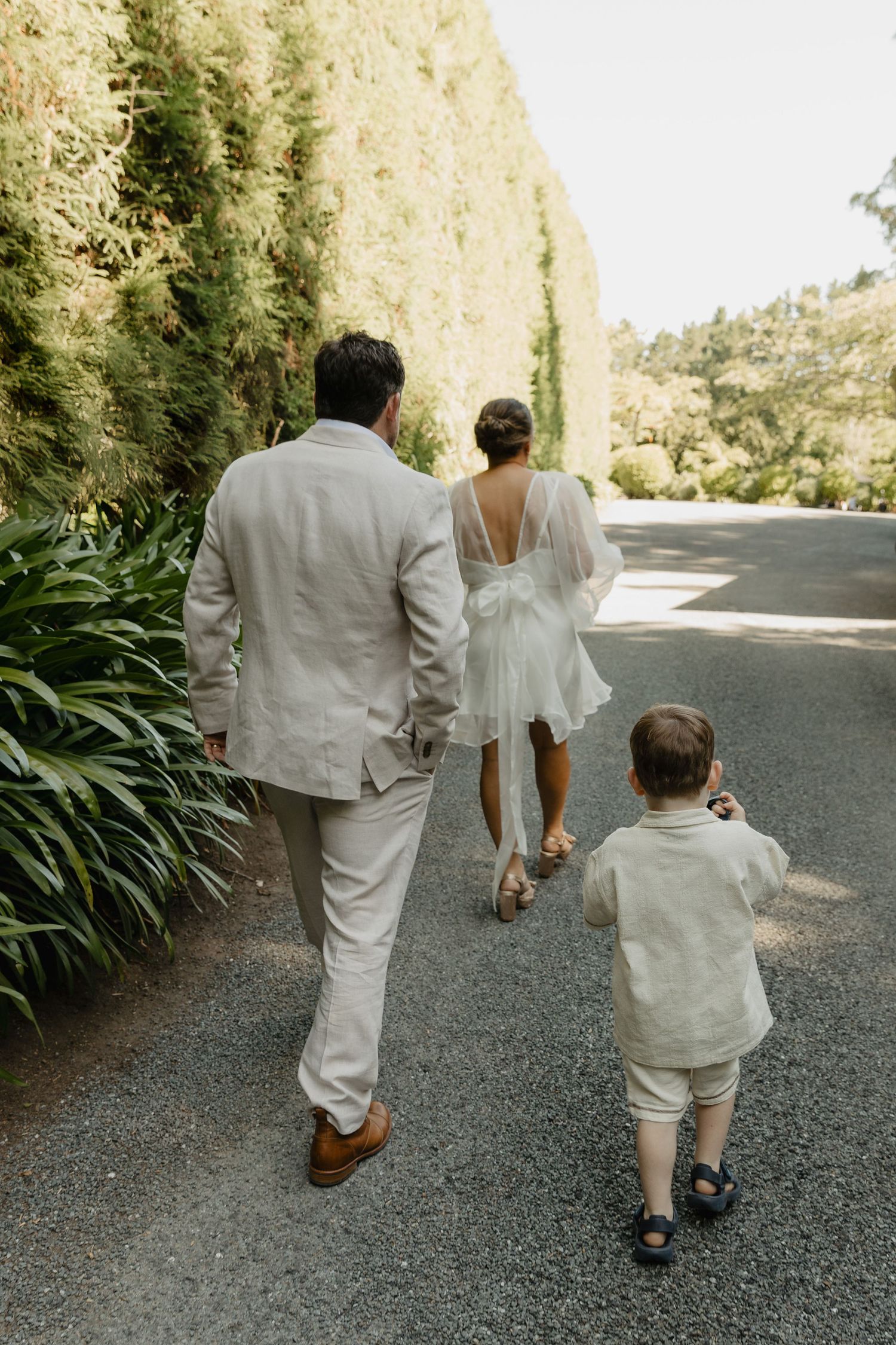 Family walking down a path lined with tall hedges, dressed in white formal attire.