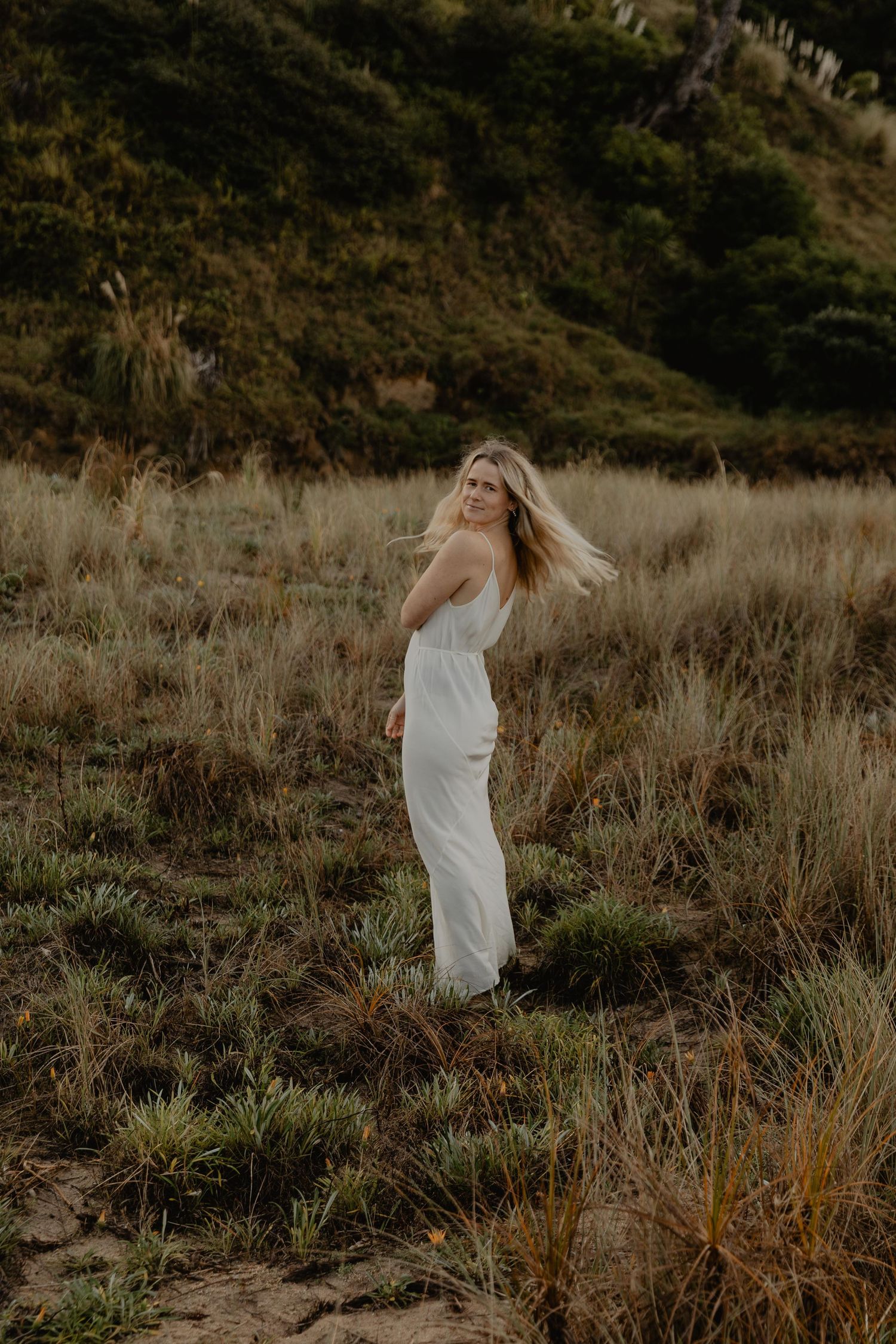 Photo of a person in white dress standing in golden field against hillside at sunset.