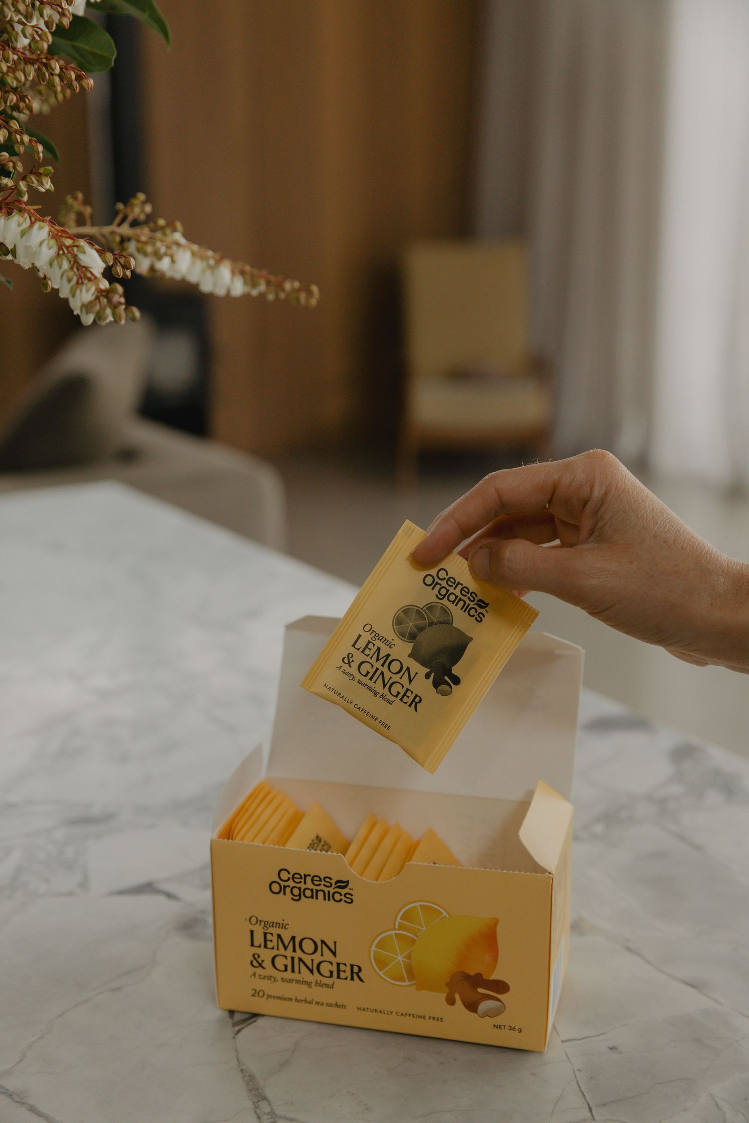 Hand removing tea bag from yellow box labeled Lemon & Ginger on marble surface.