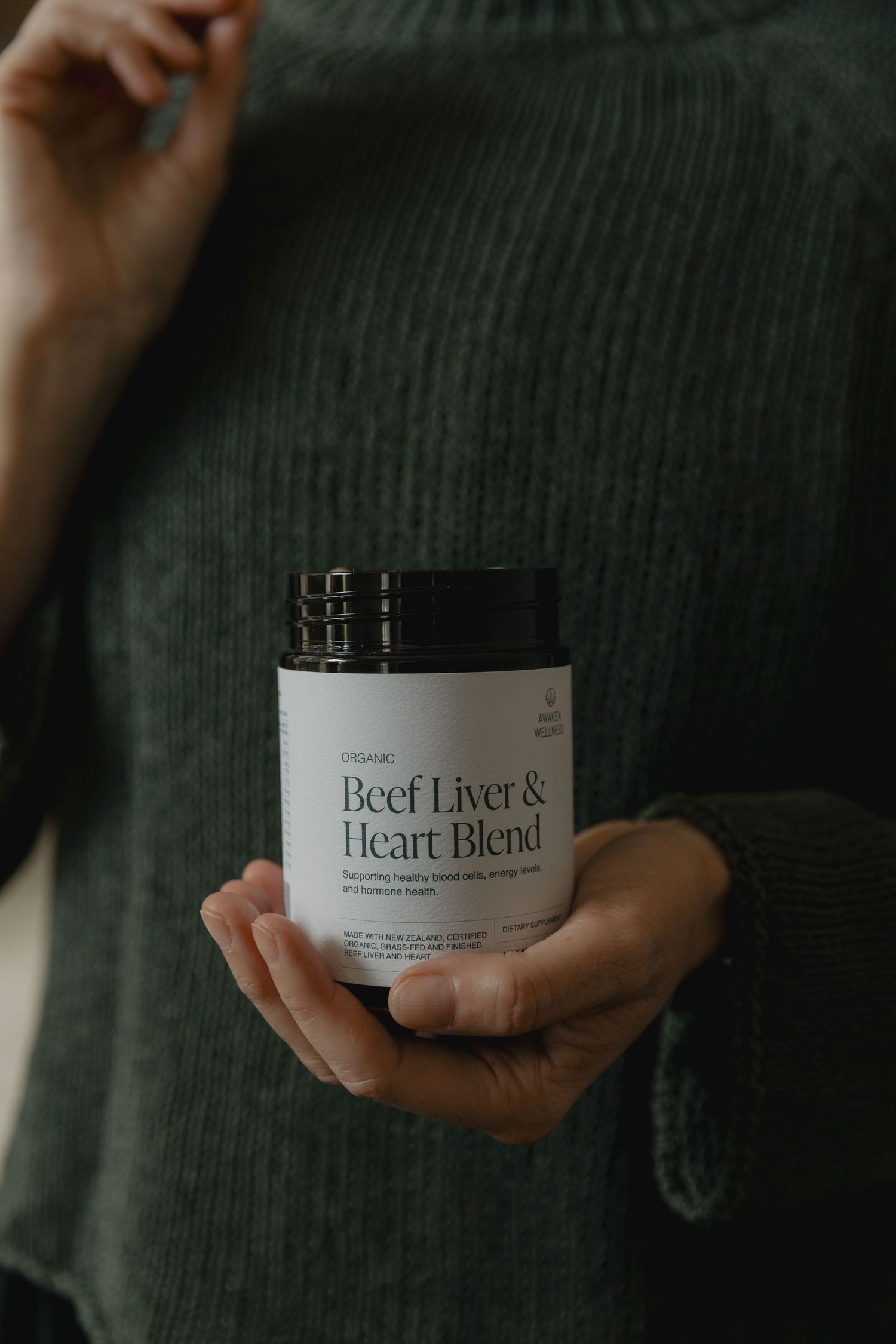 Hands holding a bottle of beef liver and heart blend supplement against a dark green sweater.