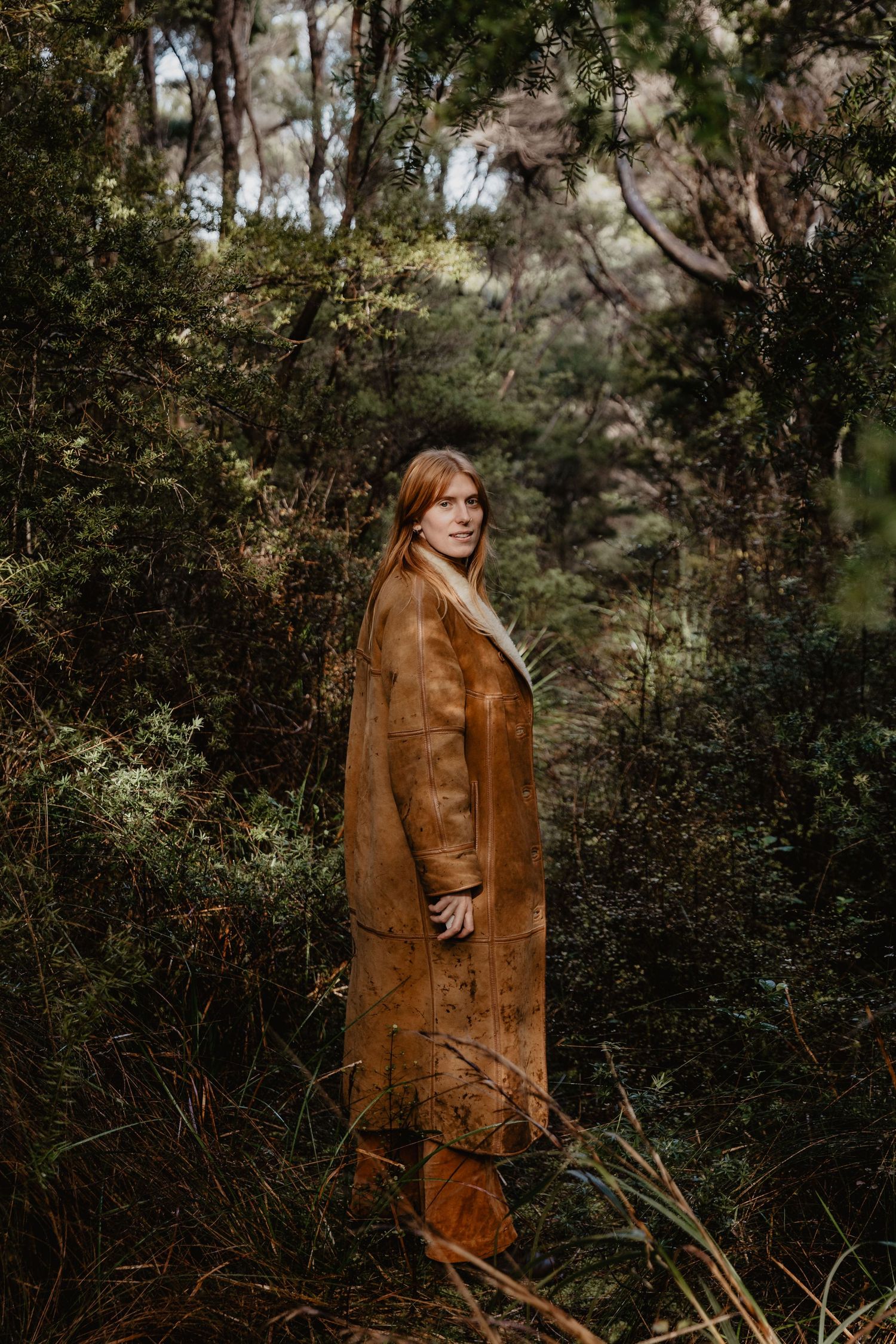 Person in brown leather jacket standing among lush forest greenery.