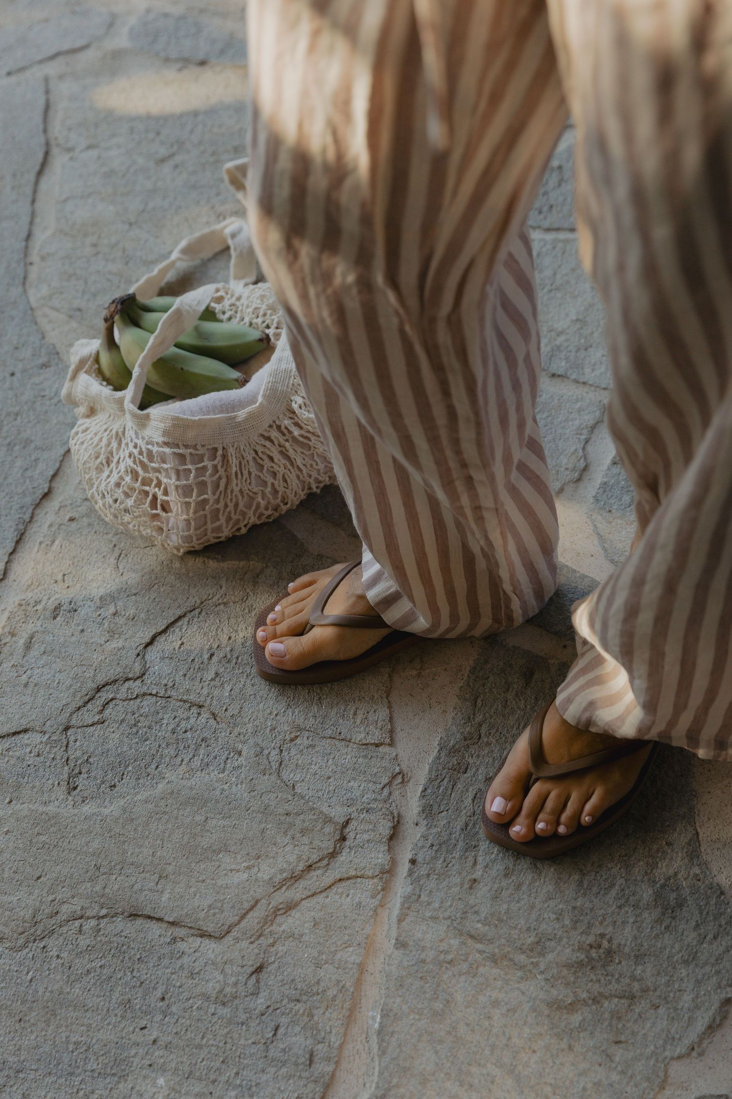 Close-up of feet wearing sandals with a flowing striped dress hem on stone ground.