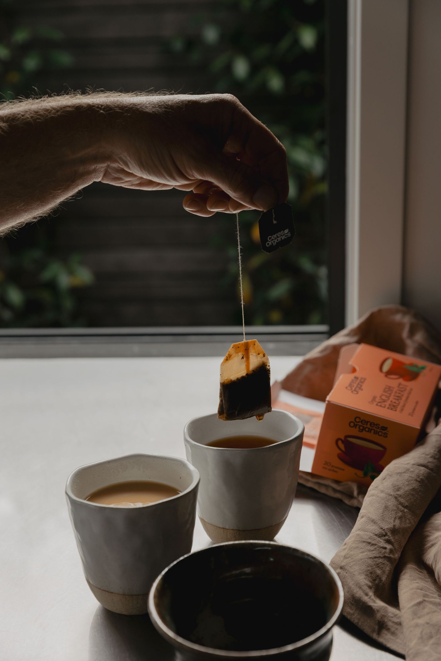 Hand dipping a tea bag into a ceramic cup with additional cups and tea packaging visible.