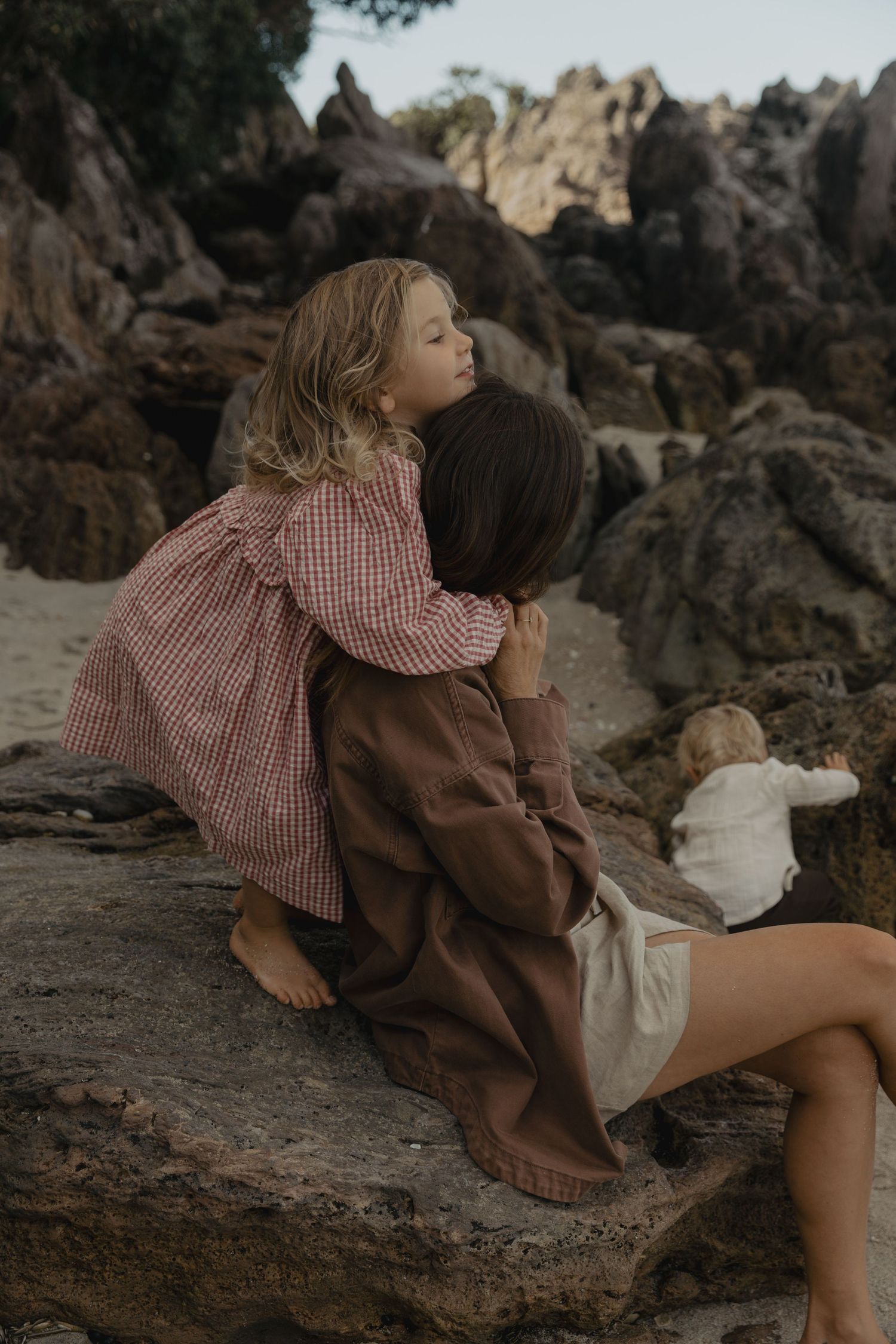 A child in a red gingham dress rests on dark coastal rocks at the beach.