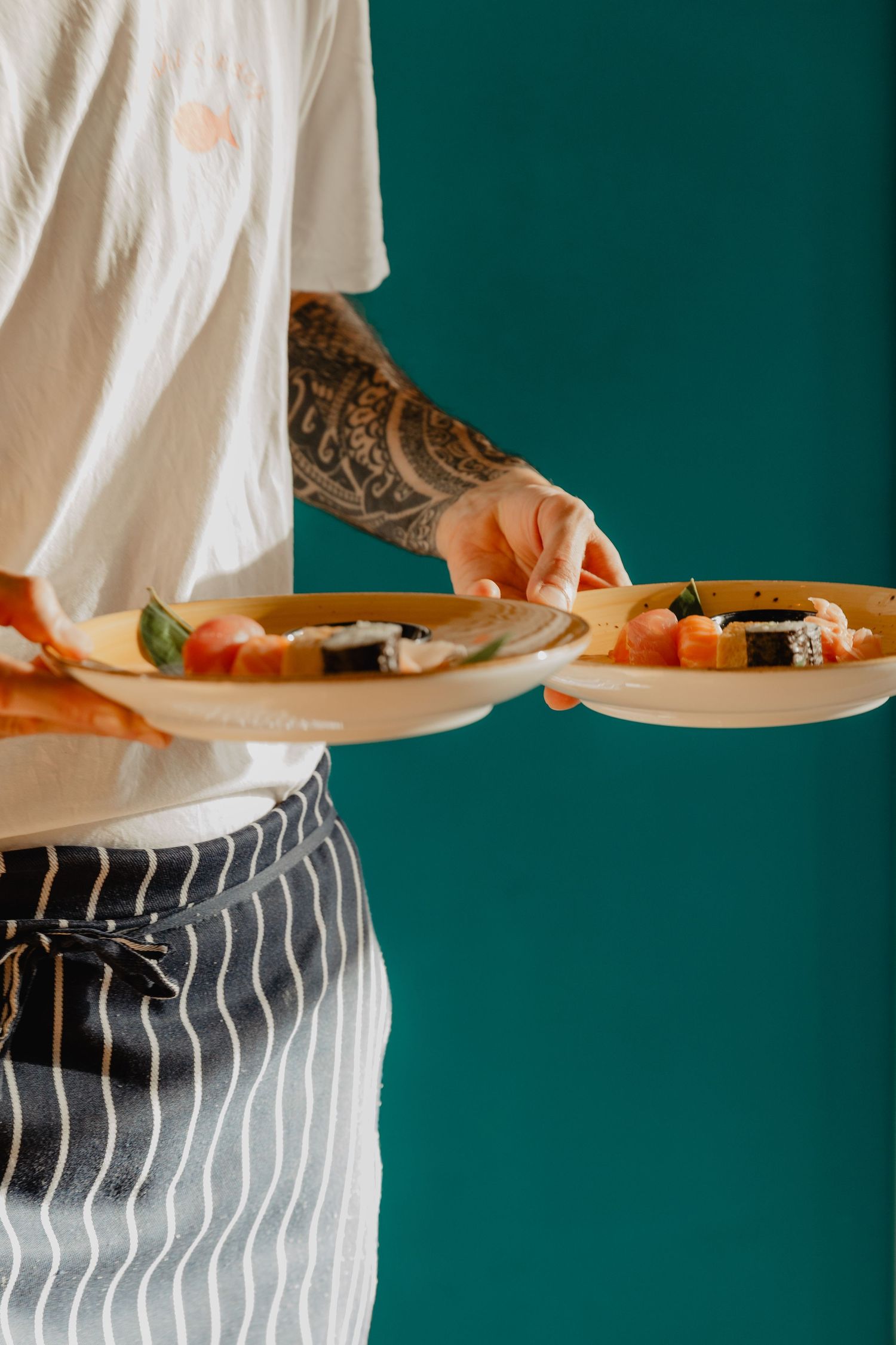 Chef in striped apron and white shirt preparing food against dark green wall.
