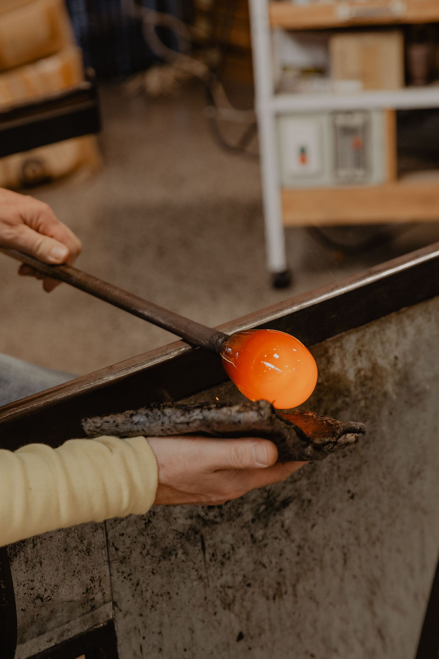 Hands shape molten glass using metallic tools in a glassblowing studio.