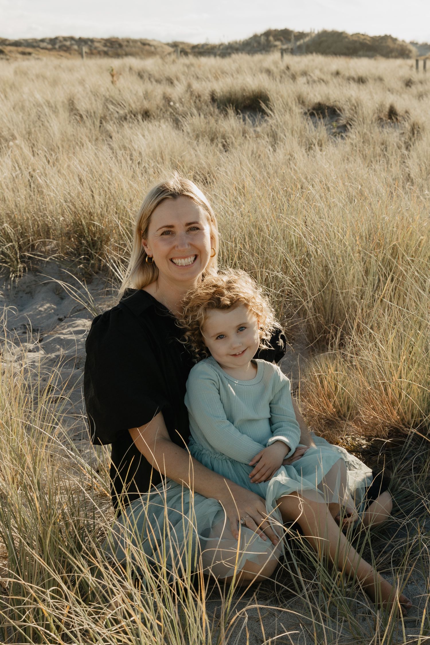 A parent and child share tender moments while sitting together in golden dry grass during sunset.