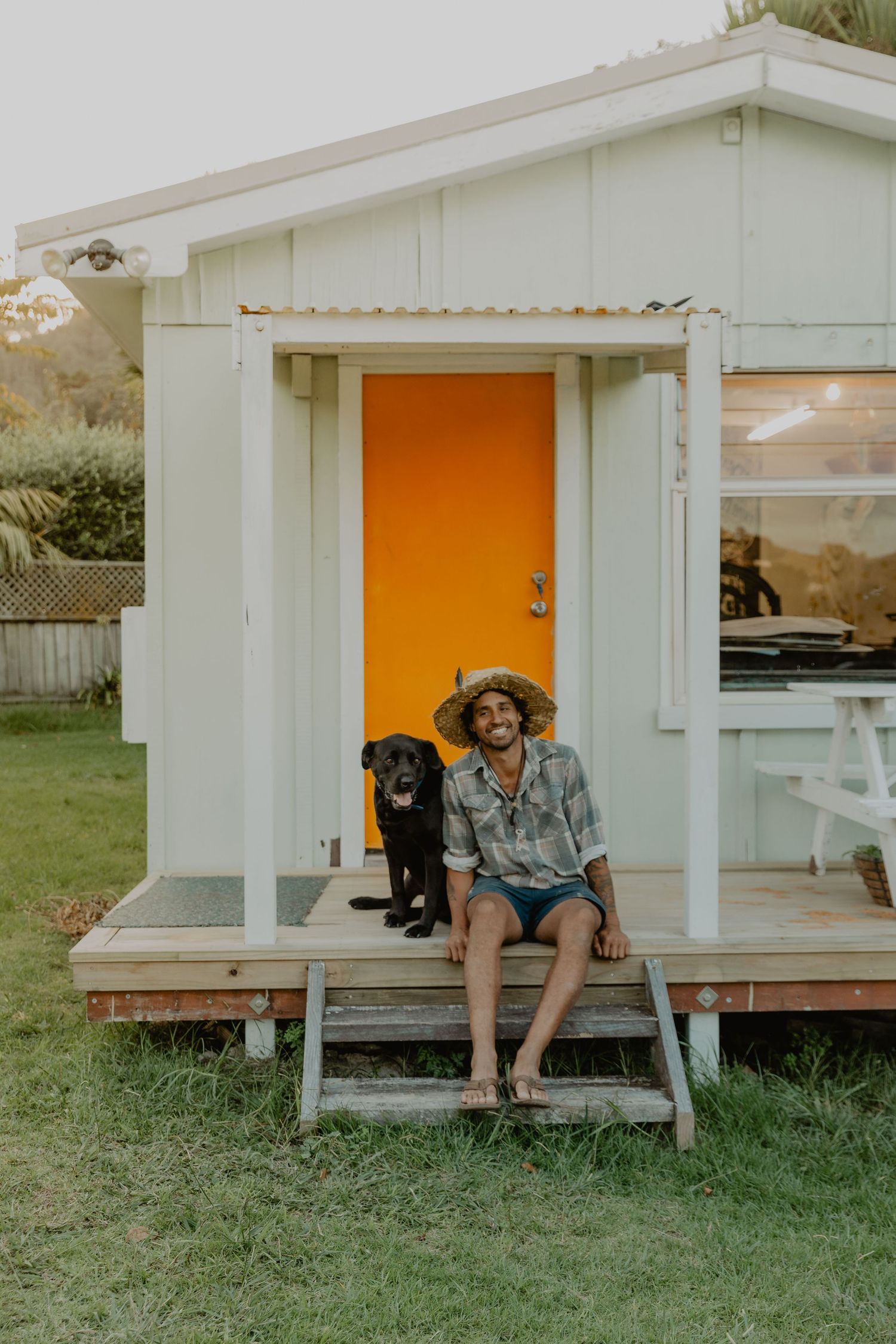 Black dog and companion sitting on porch of small white house with orange door.
