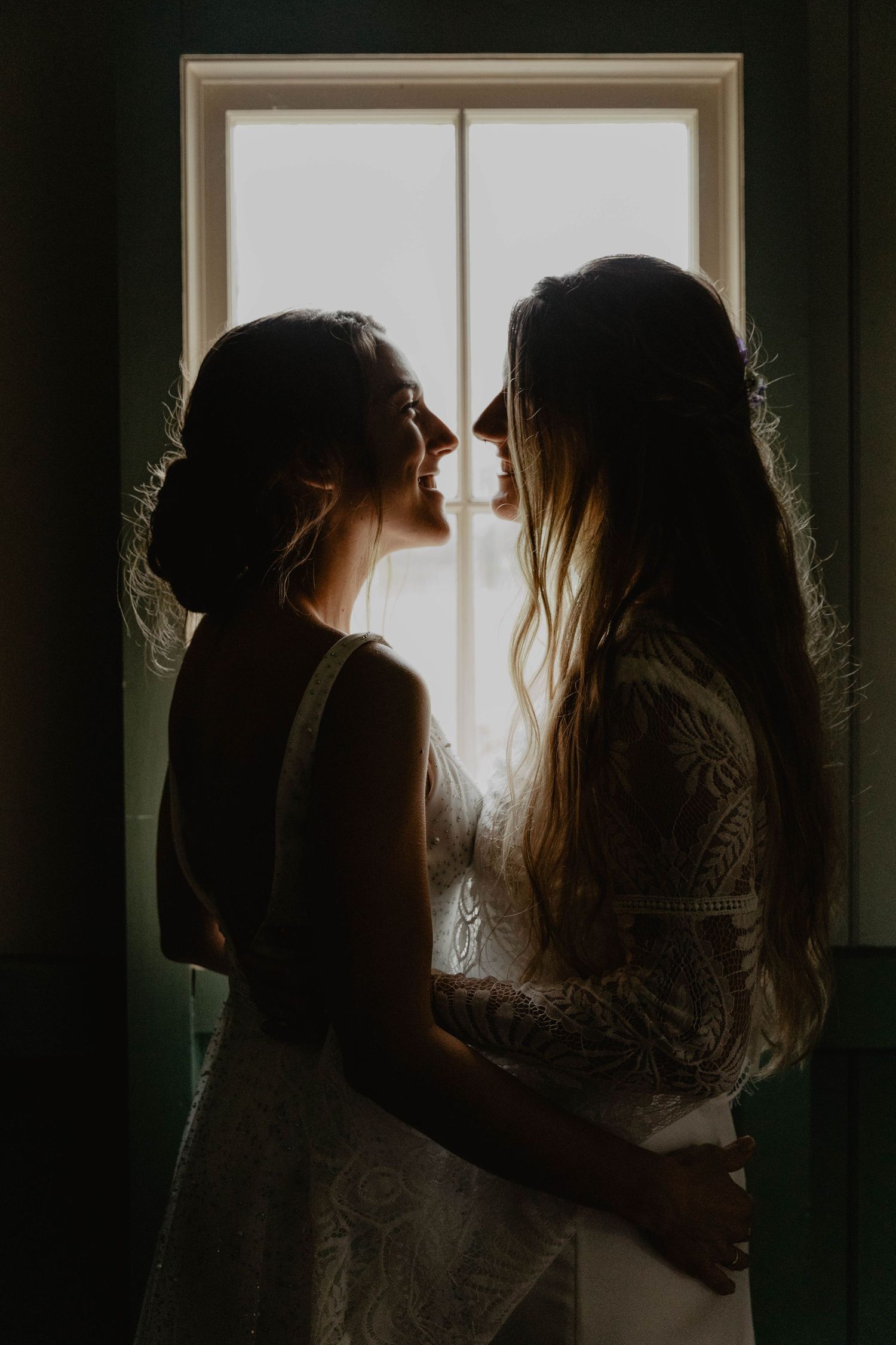 Two brides in white dresses share intimate moment by window in dramatic lighting.