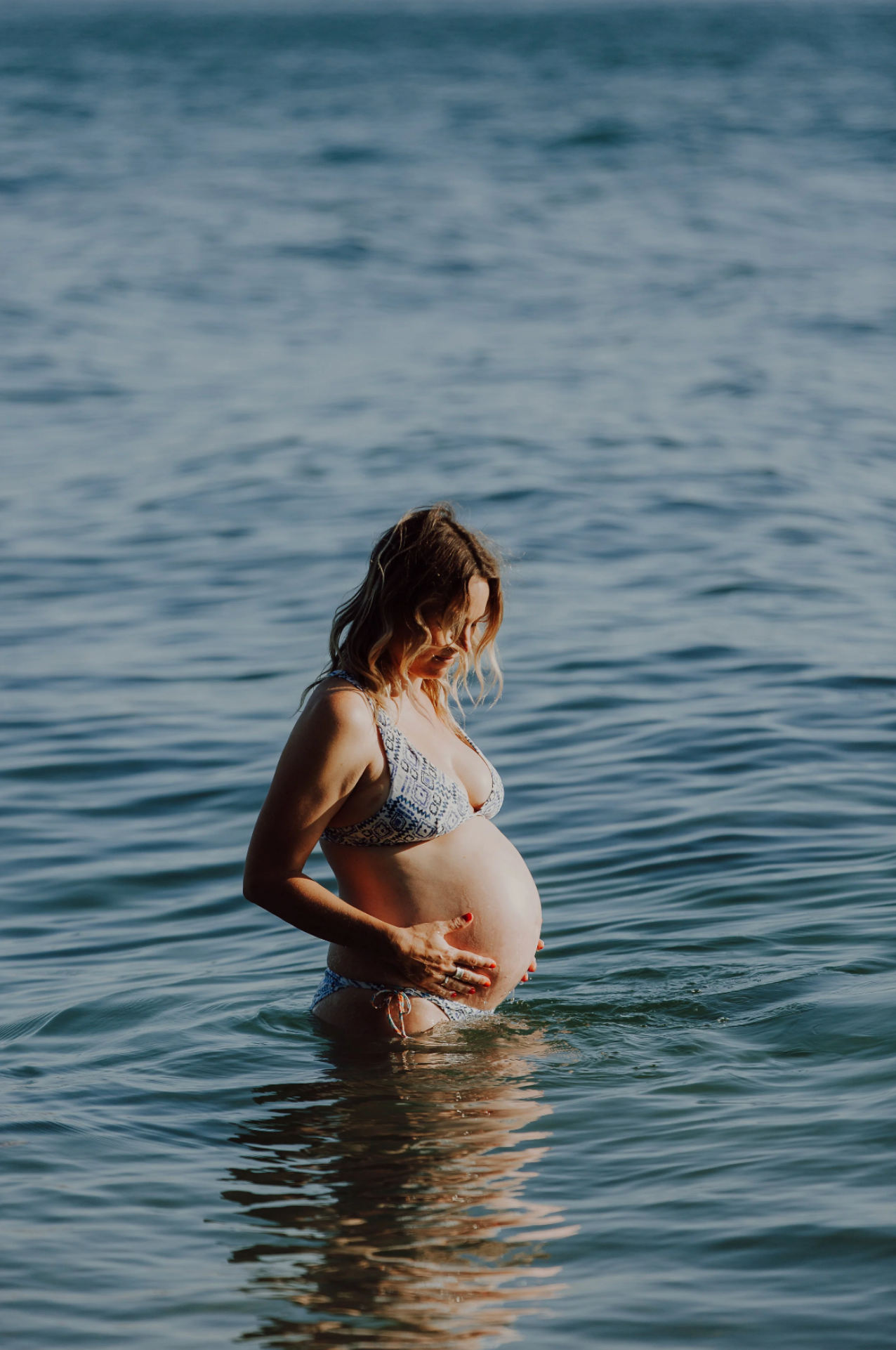 Expectant mother stands in ocean water during sunset maternity photoshoot.