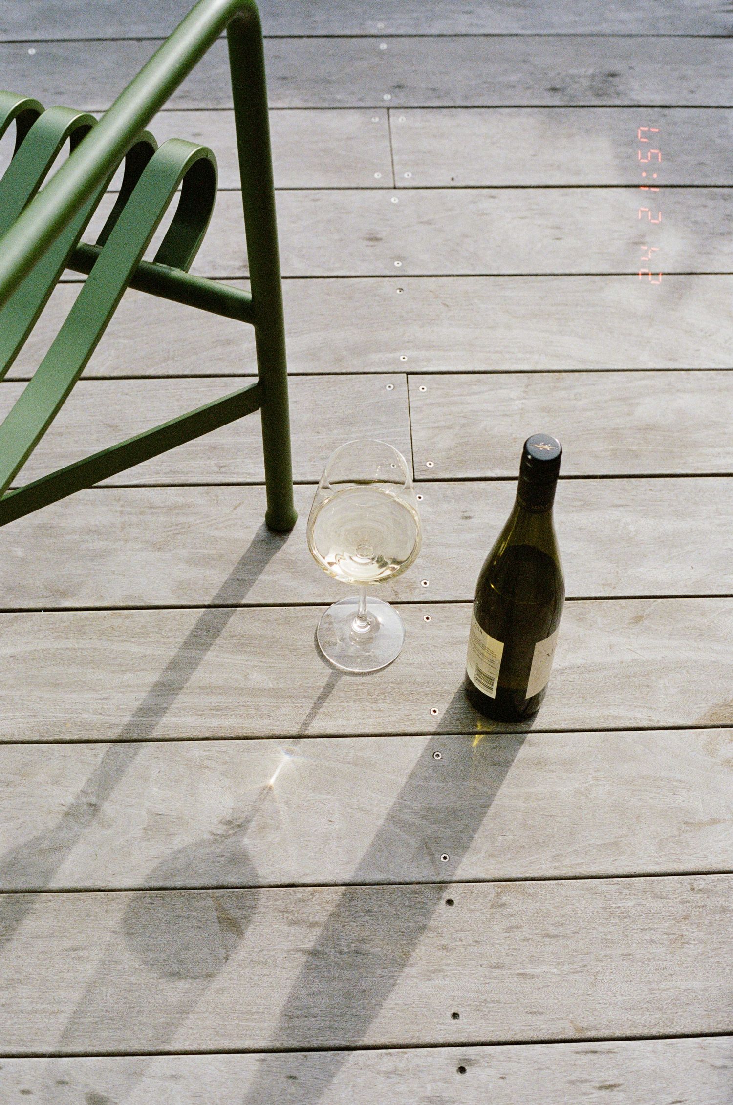 Green metal chair legs casting shadows on a wooden deck with wine glasses.