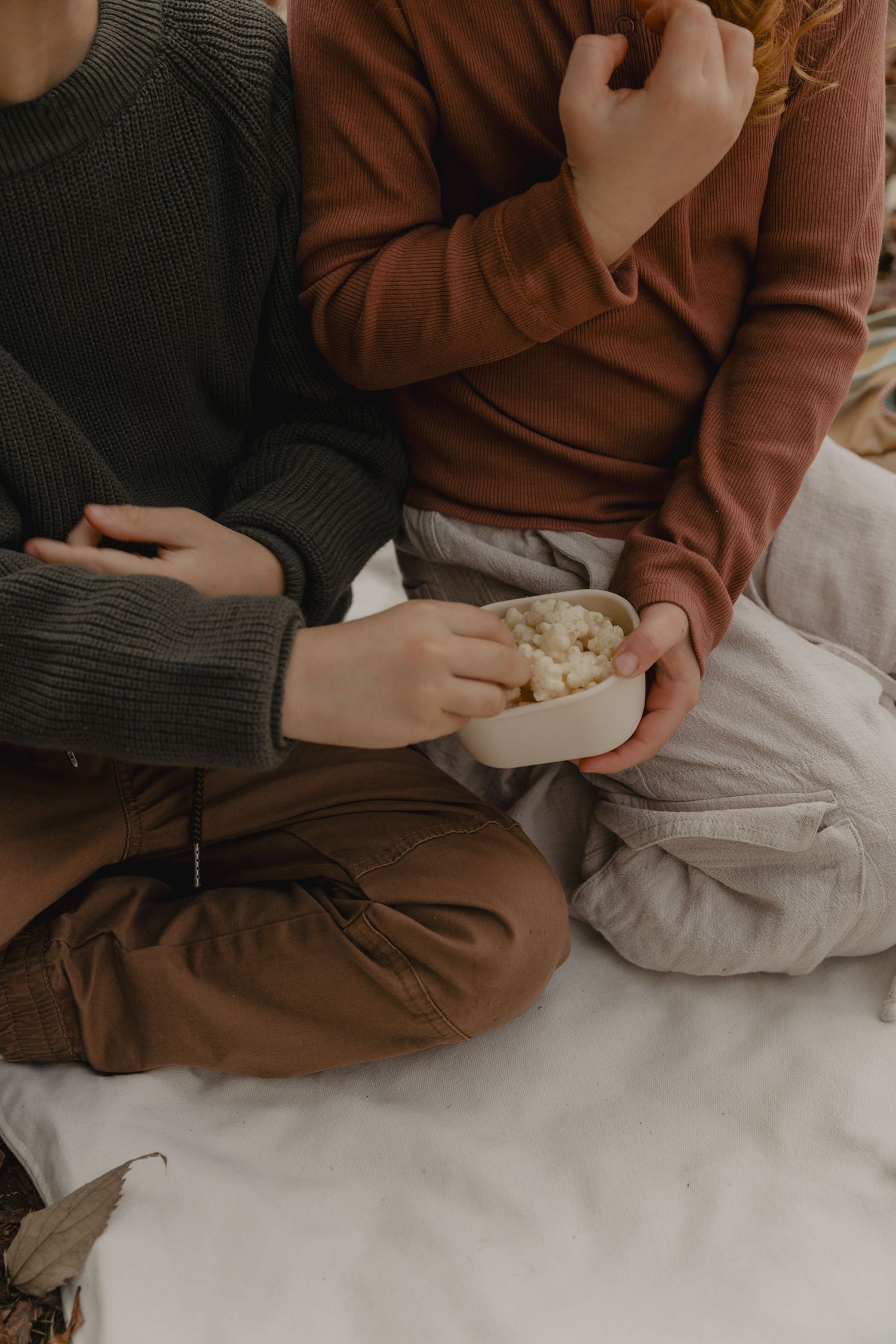 People in cozy sweaters relaxing on white bedding with snacks.