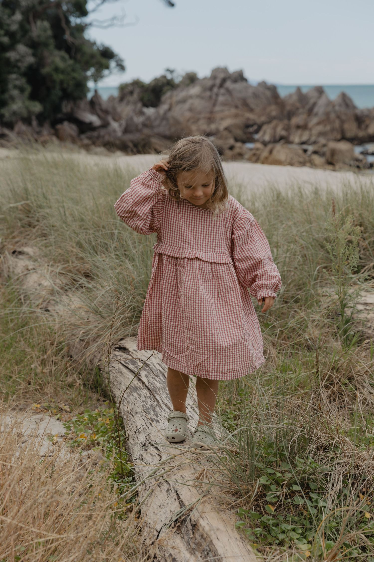 Child in gingham dress playing in tall grass near rocky coastline.
