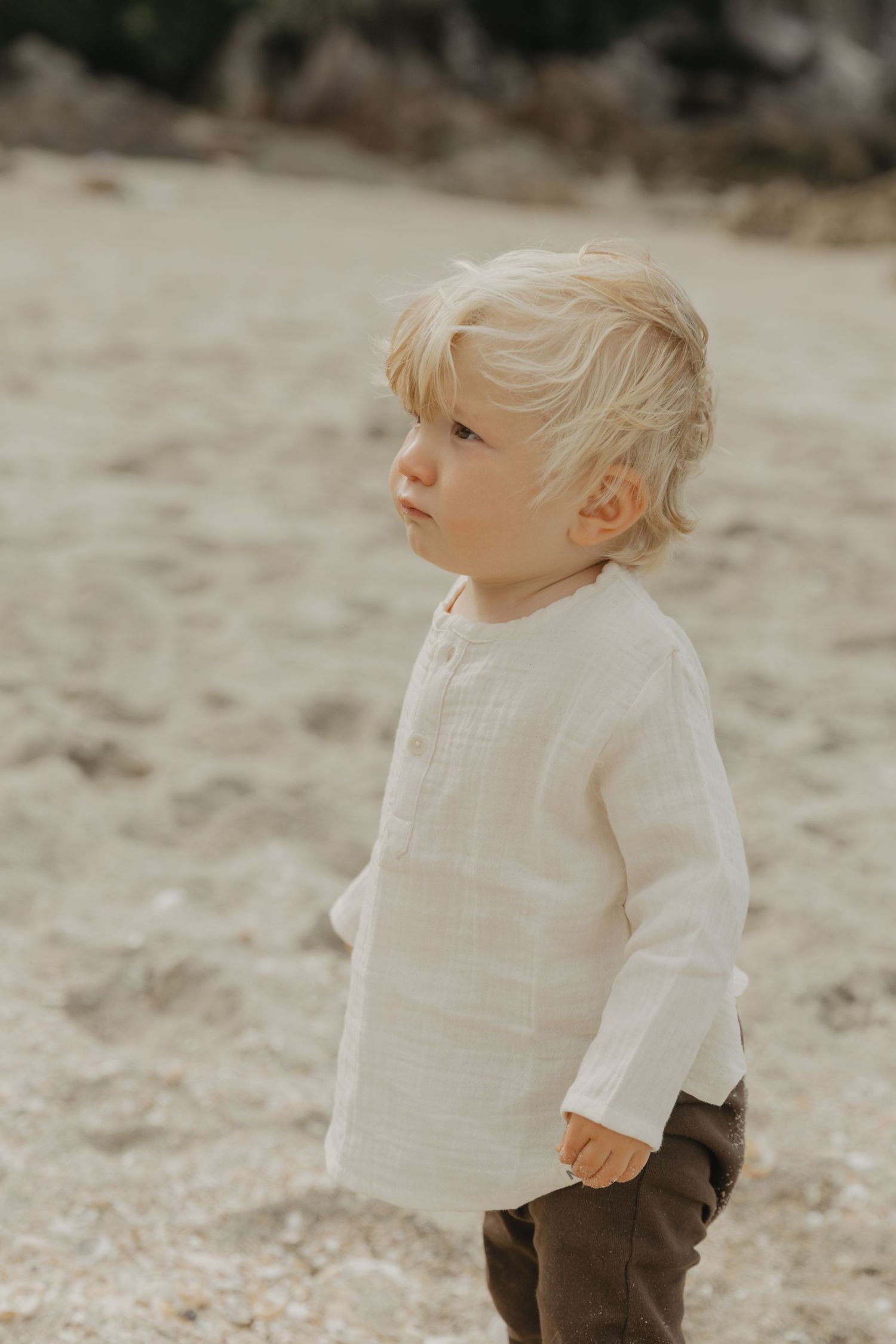 A young child in a white linen shirt and brown pants stands on a sandy beach.
