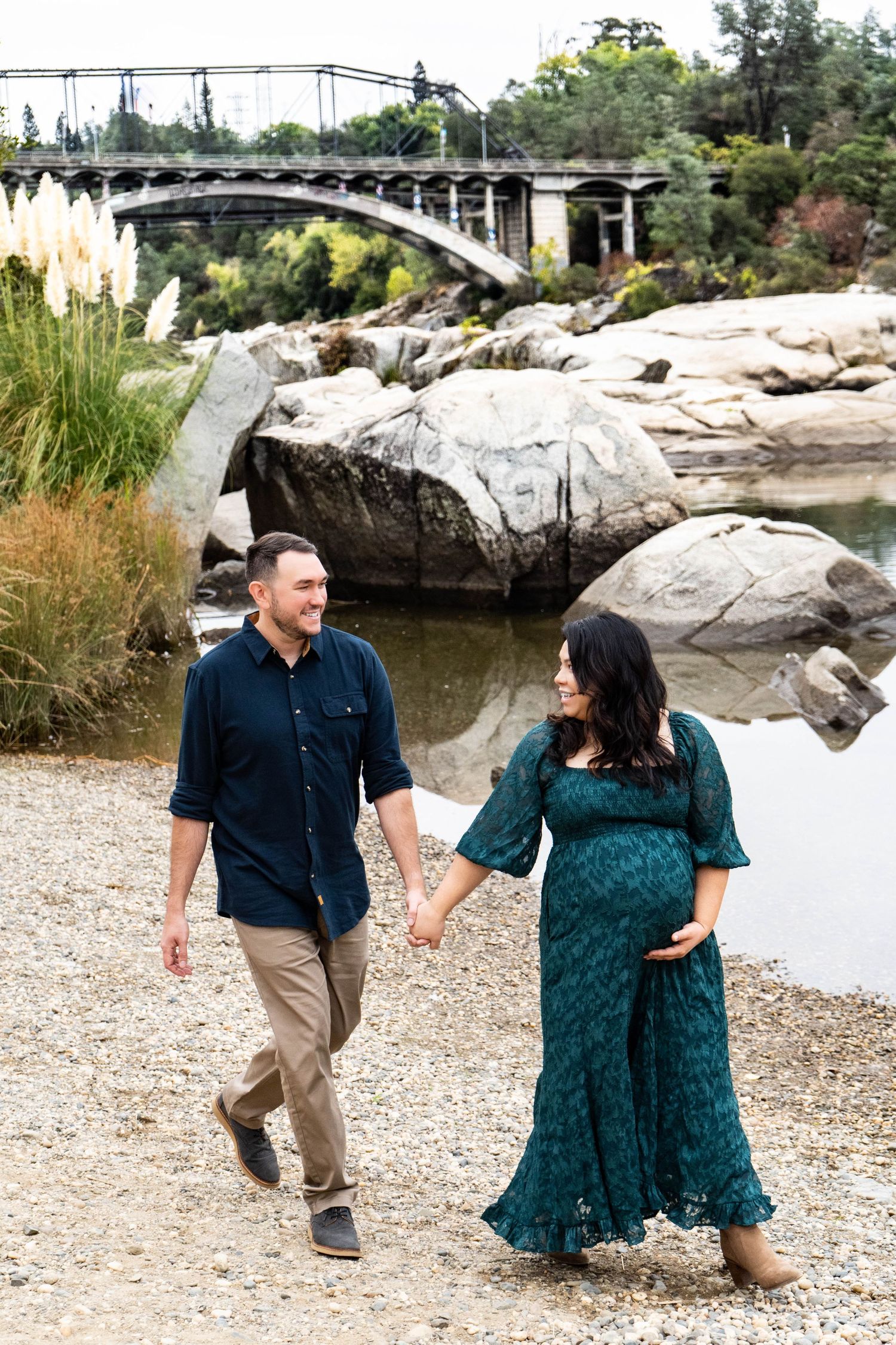 A couple walks hand in hand along a rocky riverbank with a bridge visible in the background.