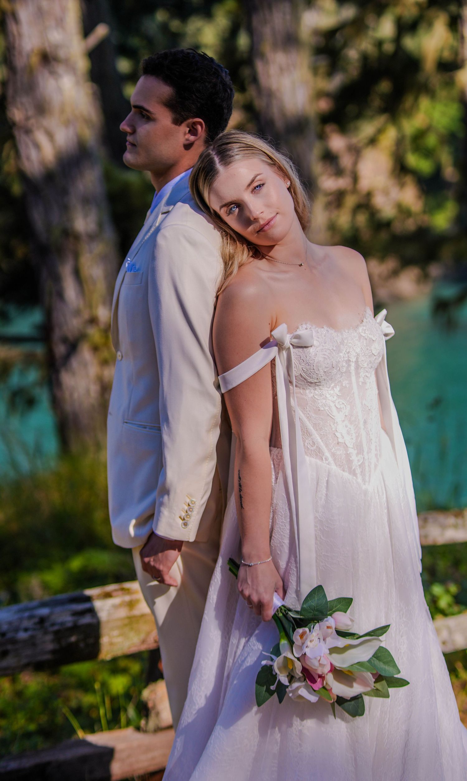 Bride in white gown holds bouquet leaning on groom in cream suit outdoors at a romantic wedding ceremony.