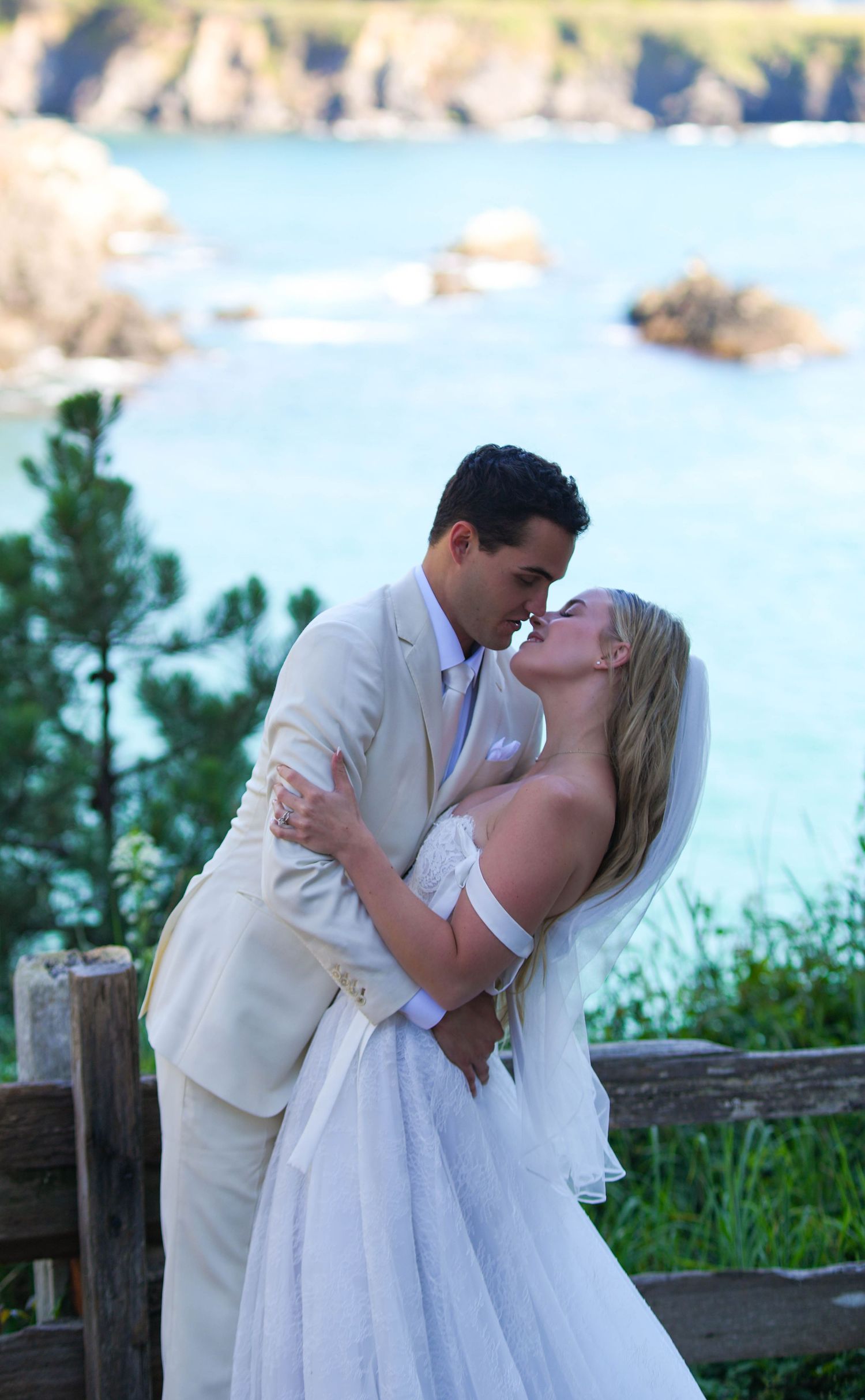 Bride and groom share a romantic kiss at coastal wedding venue with turquoise ocean and rocky shoreline backdrop.