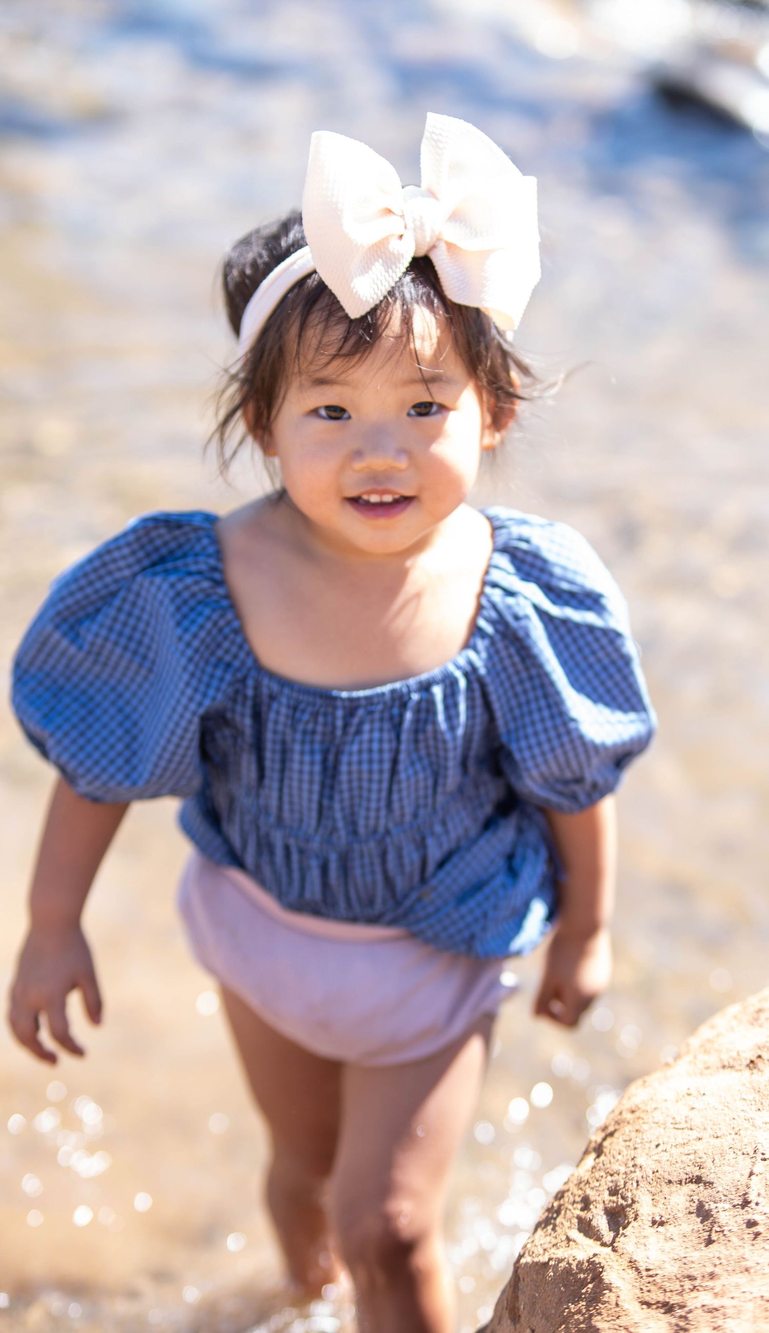 Small child in blue gingham dress and white bow playing on sandy beach.