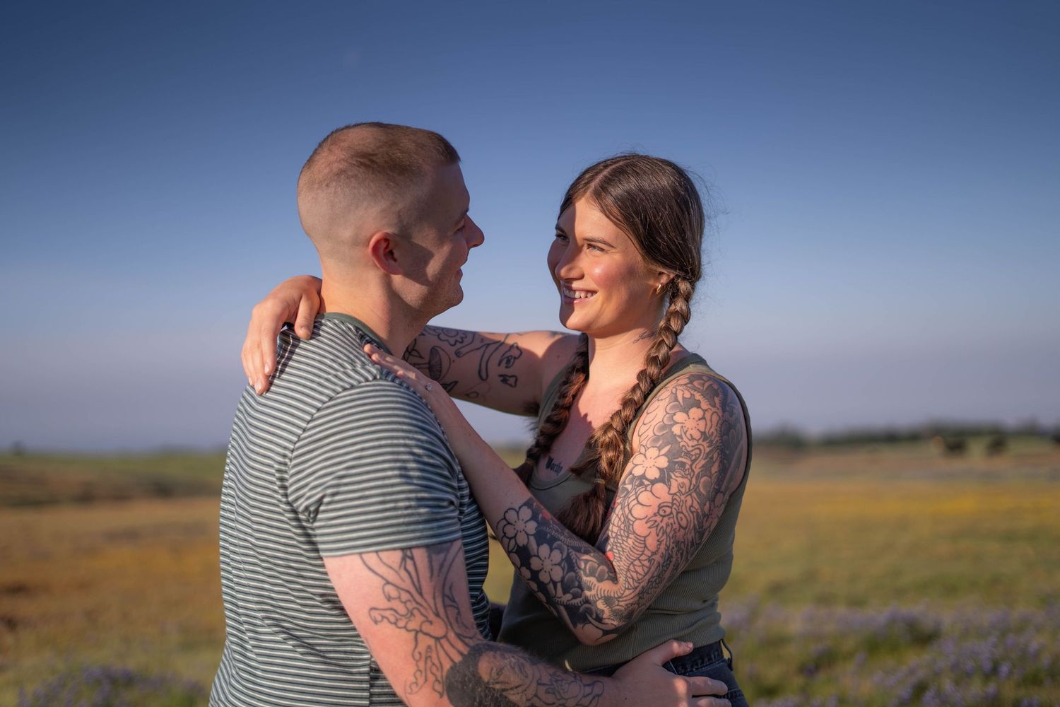 Couple with tattoos embracing and smiling at each other in a sunny field with wildflowers and clear blue sky.
