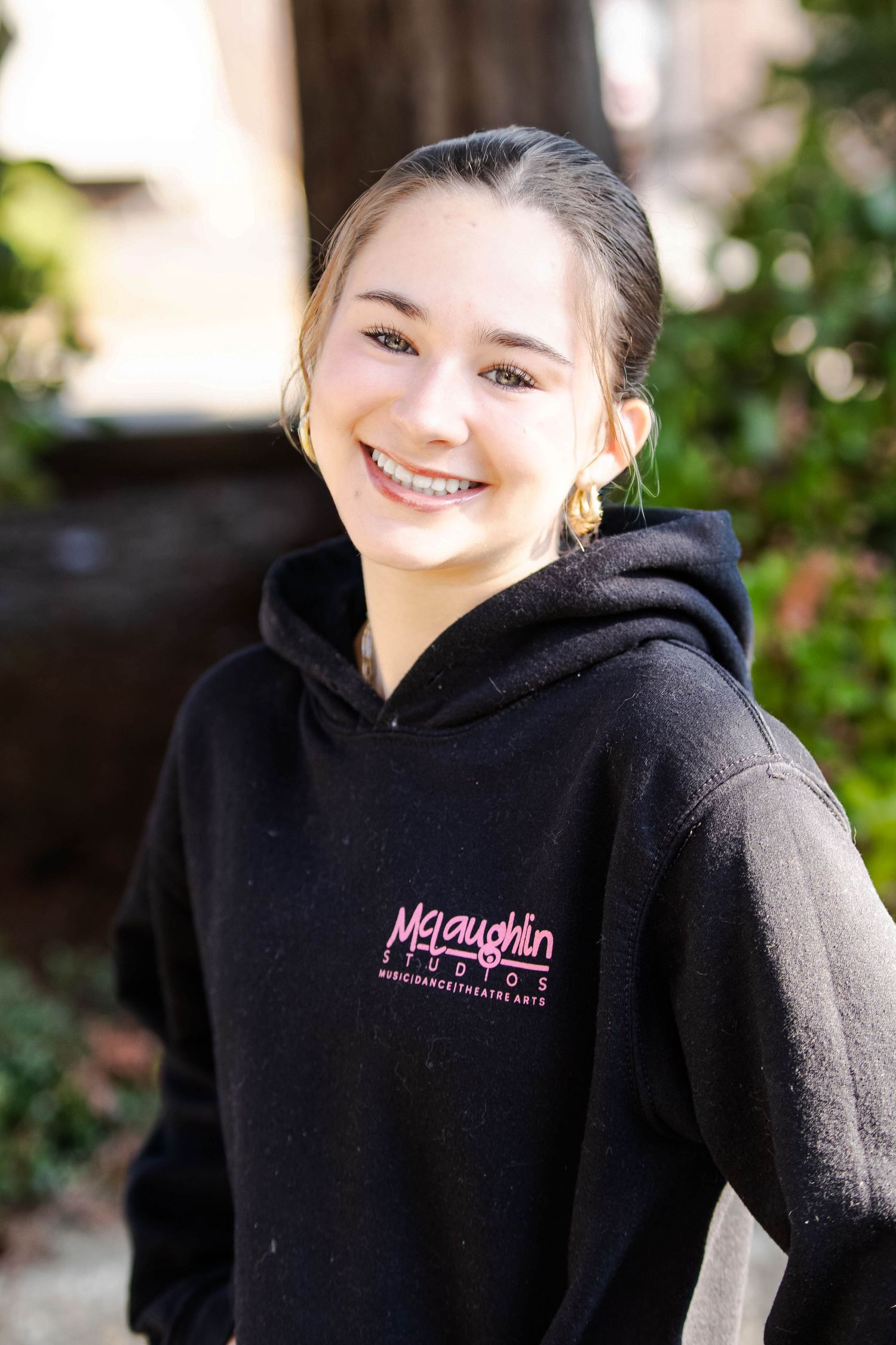 Smiling teenage girl wearing a black McLaughlin Studios hoodie outdoors, with braces and hair pulled back.