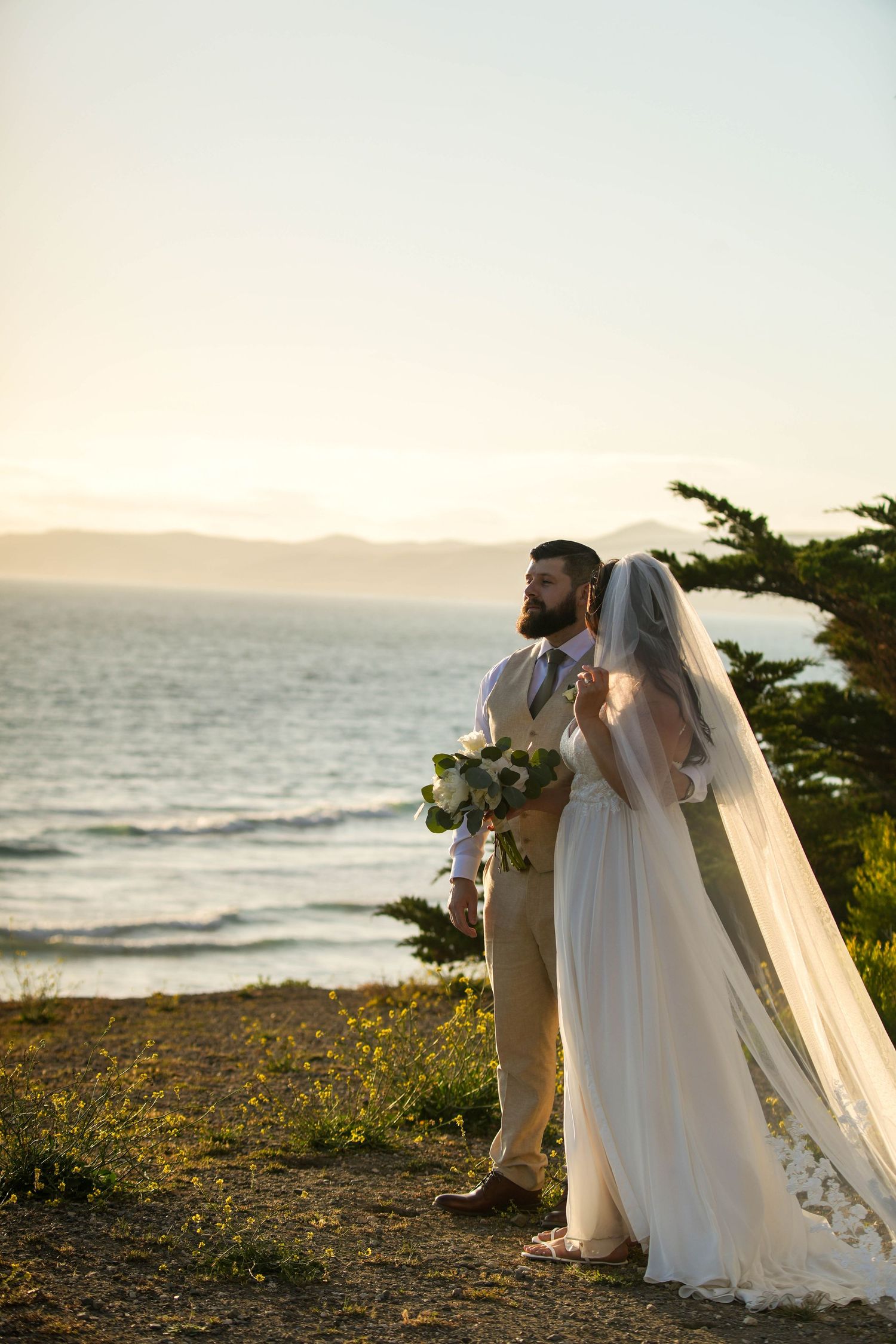 Bride and groom stand together overlooking the ocean at sunset, bride wearing a flowing veil and gown holding a bouquet.
