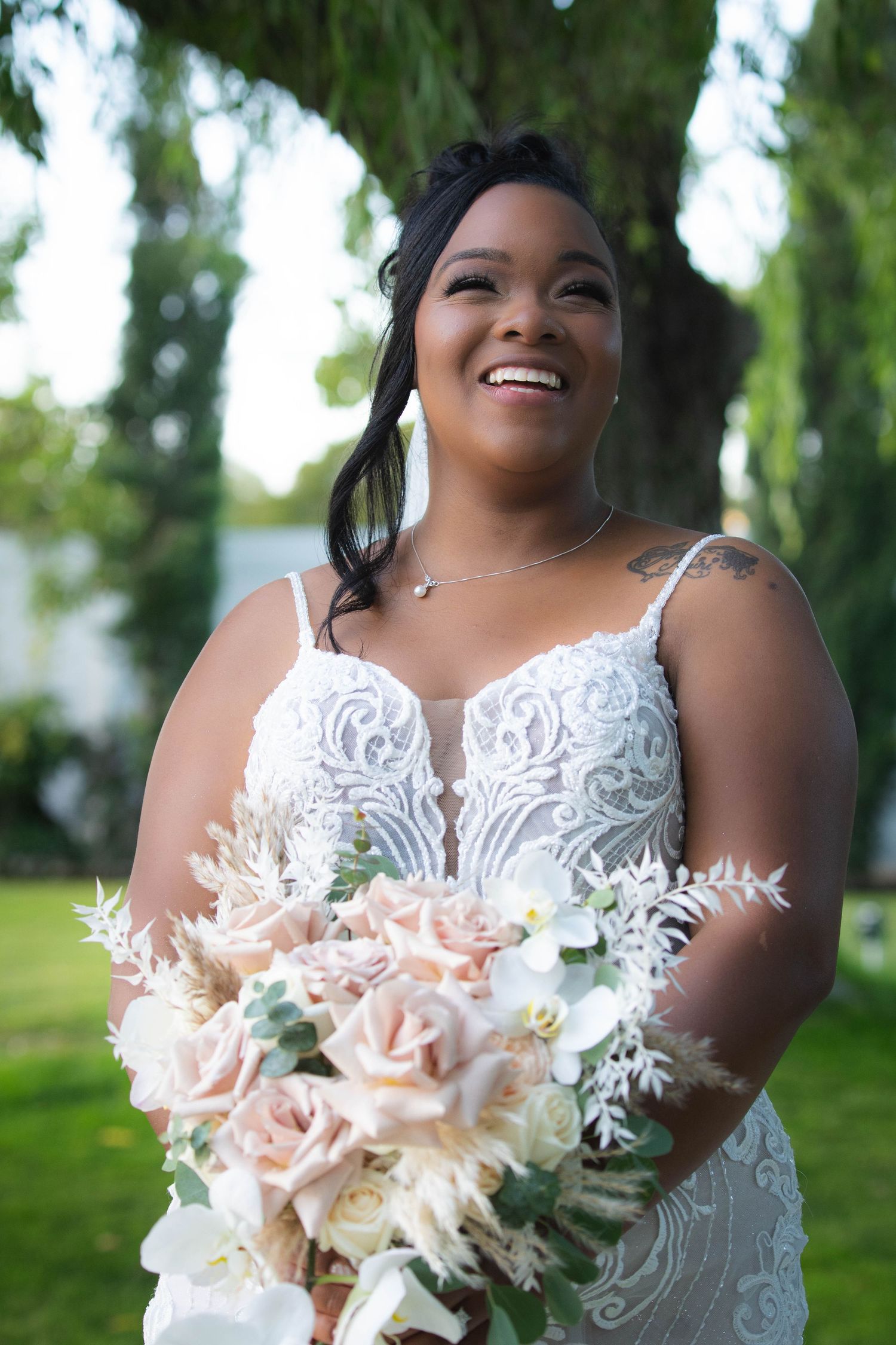 Joyful bride in white lace gown holding a blush pink and white bouquet against a green outdoor background.