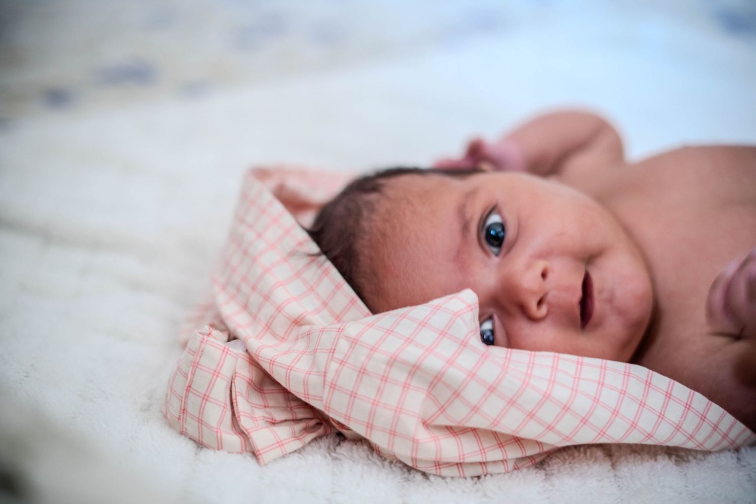Newborn baby wrapped in a pink checkered blanket lying on a white surface with bright blue eyes.