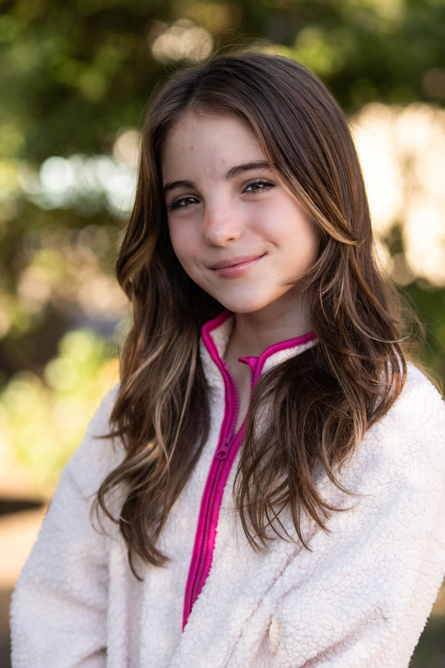 Smiling young girl with long brown hair wearing a white and pink zip-up fleece jacket outdoors.