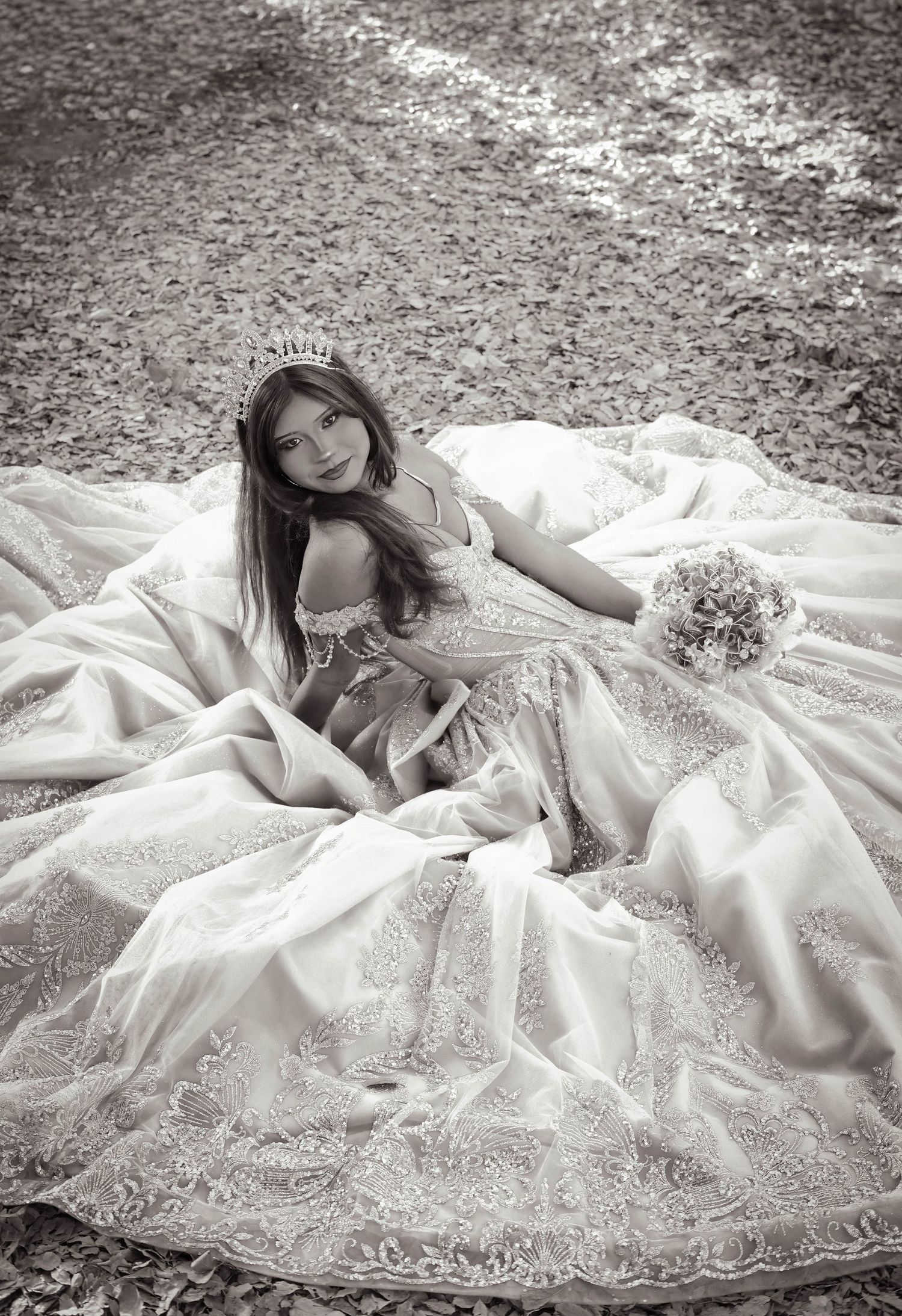 Black and white photo of a young girl in a white lace gown and tiara, seated outdoors holding a bouquet.