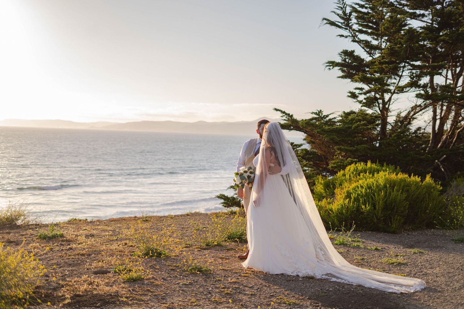 Bride in white gown and veil stands on coastal bluff overlooking ocean at golden sunset near cypress trees.