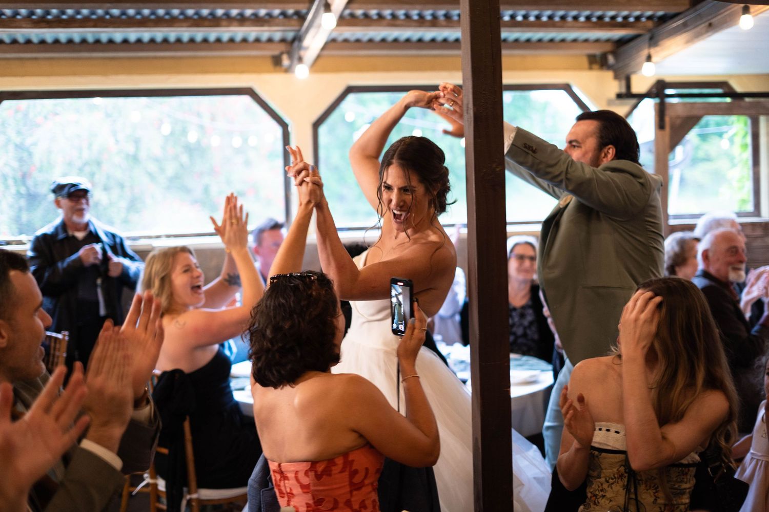 Wedding guests dancing and celebrating with raised arms under string lights at an indoor reception.
