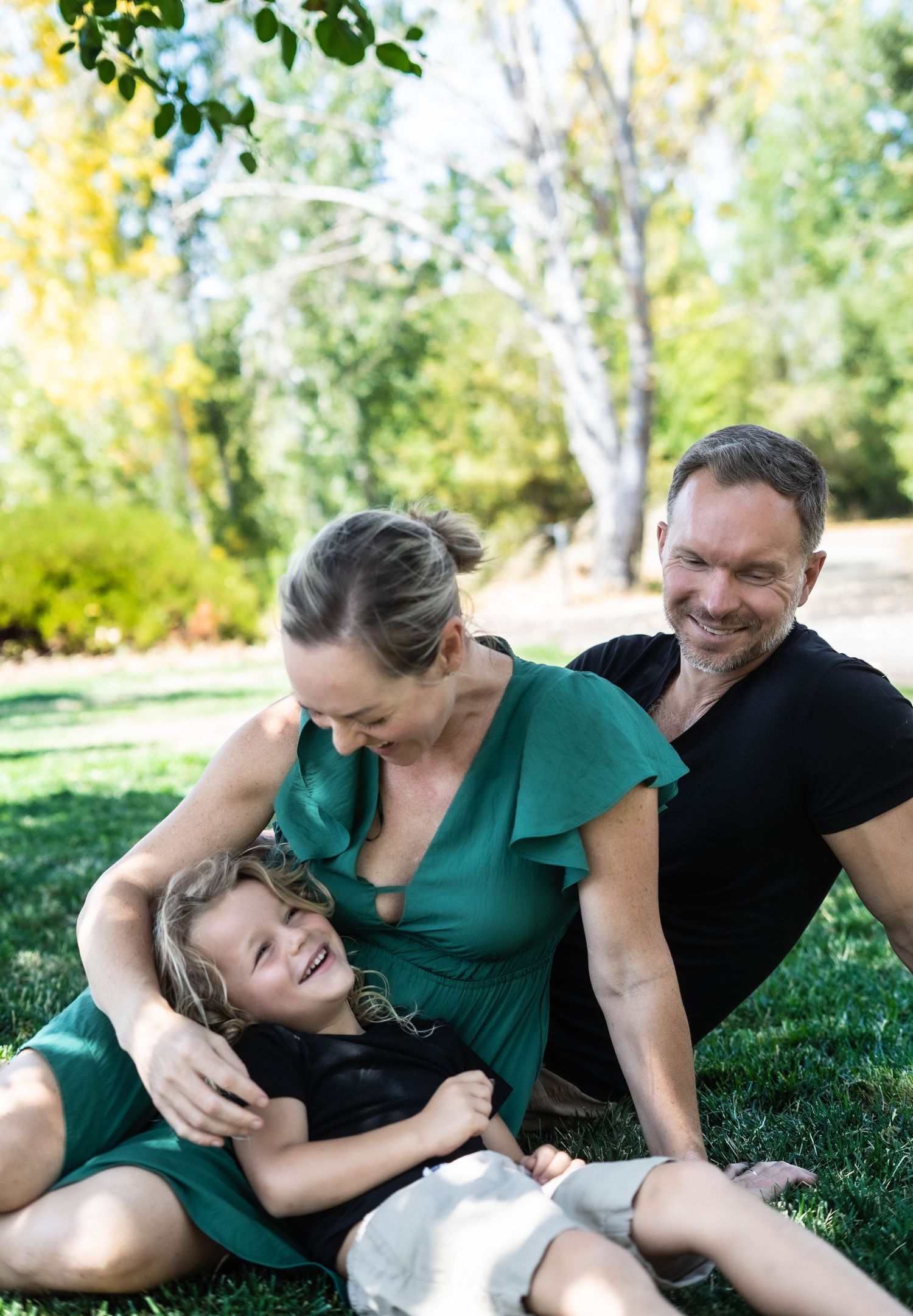 Playful family interaction on grass under trees on a sunny day.