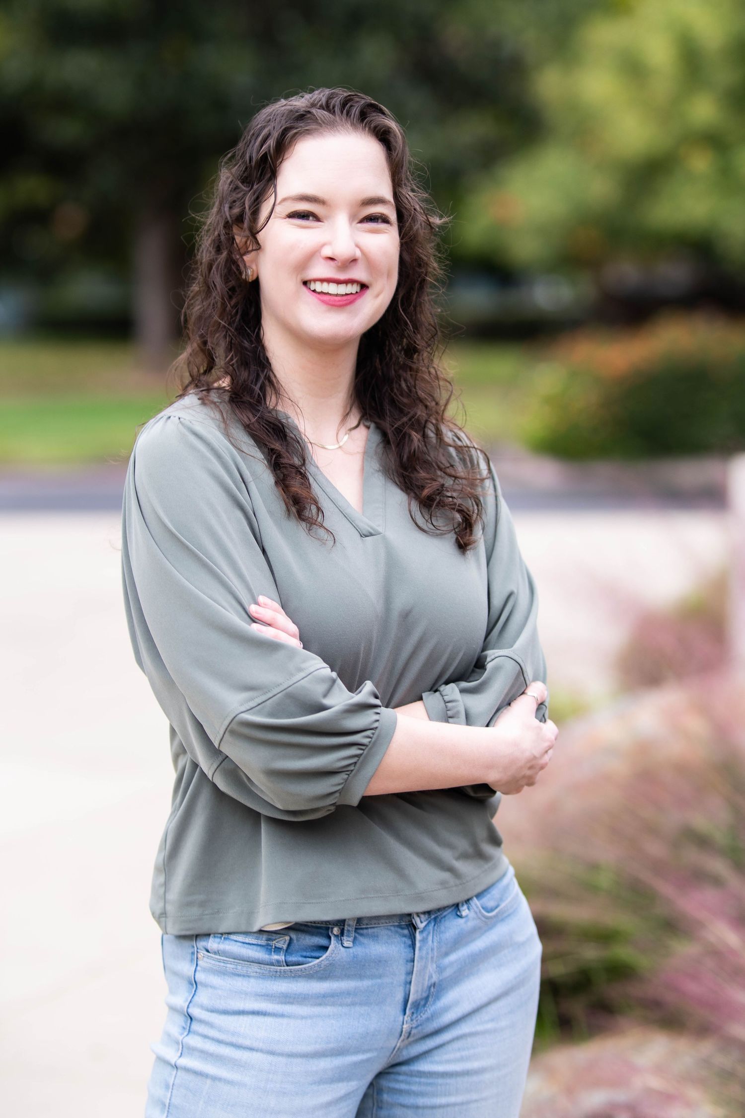 A smiling young woman with curly brown hair stands outdoors with arms crossed, wearing a sage green blouse and jeans.