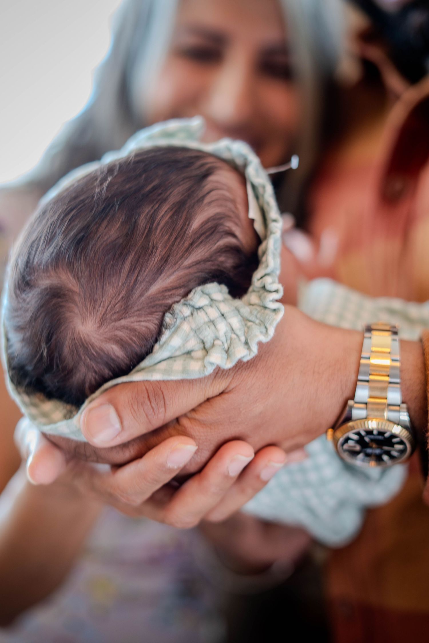 Woman with gray hair wearing a Rolex watch tenderly cradling a newborn baby wrapped in a plaid blanket.