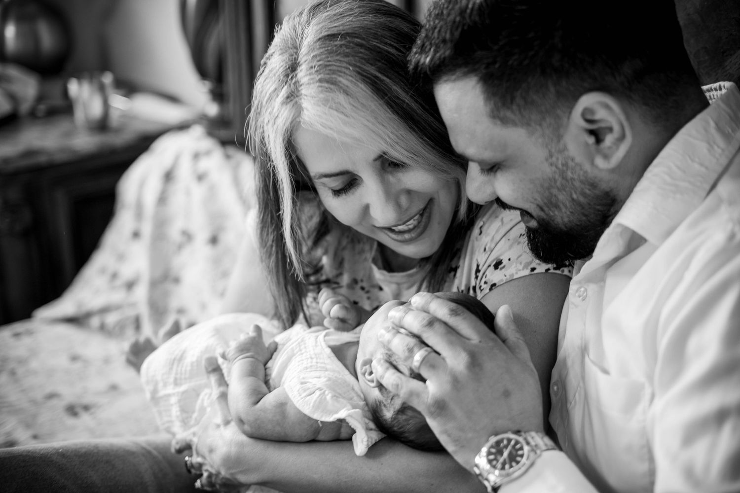 Black and white photo of smiling parents tenderly holding their newborn baby together in a heartwarming family portrait.