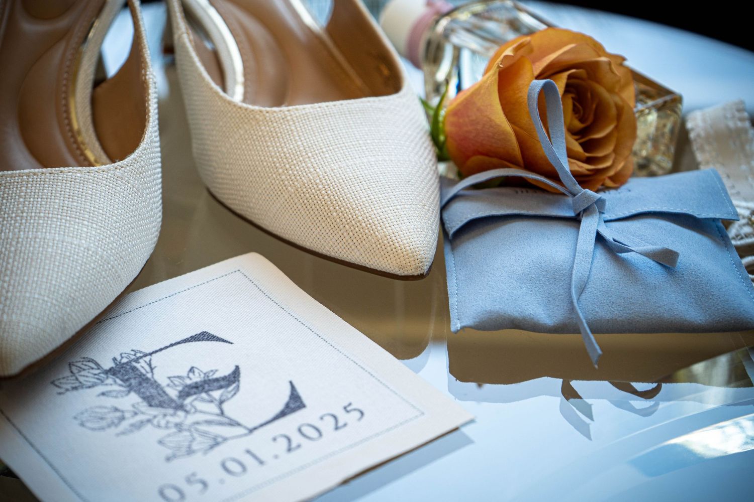 Elegant wedding flat lay with ivory heels, blue gift box, orange rose, and save the date card reading 05.01.2025.