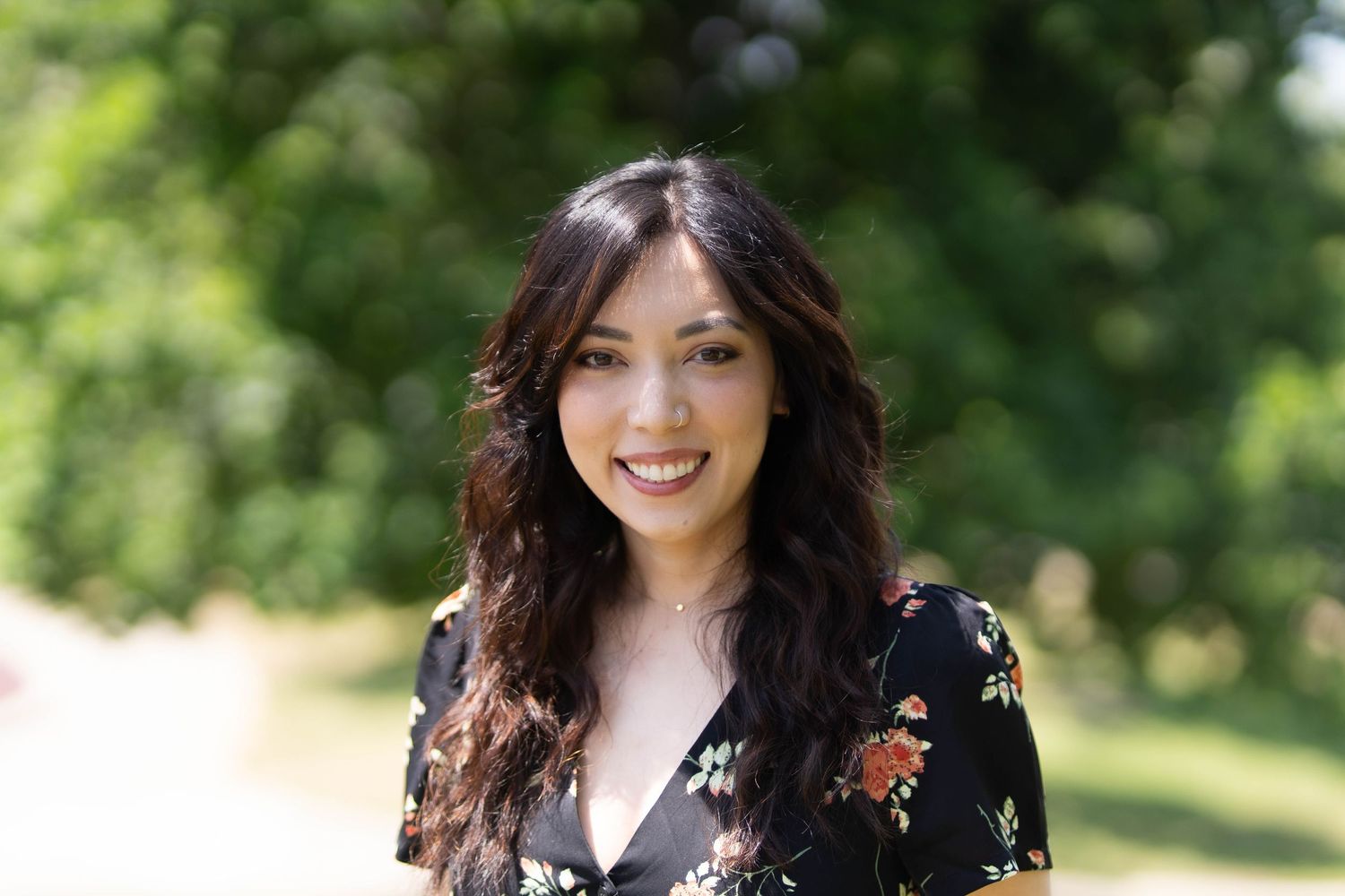 Woman in black floral dress posing outdoors in natural sunlight.