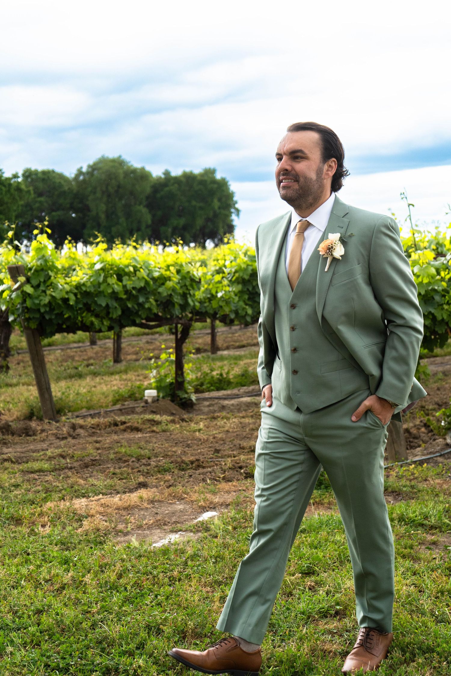 Groom in sage green suit poses confidently in vineyard setting.