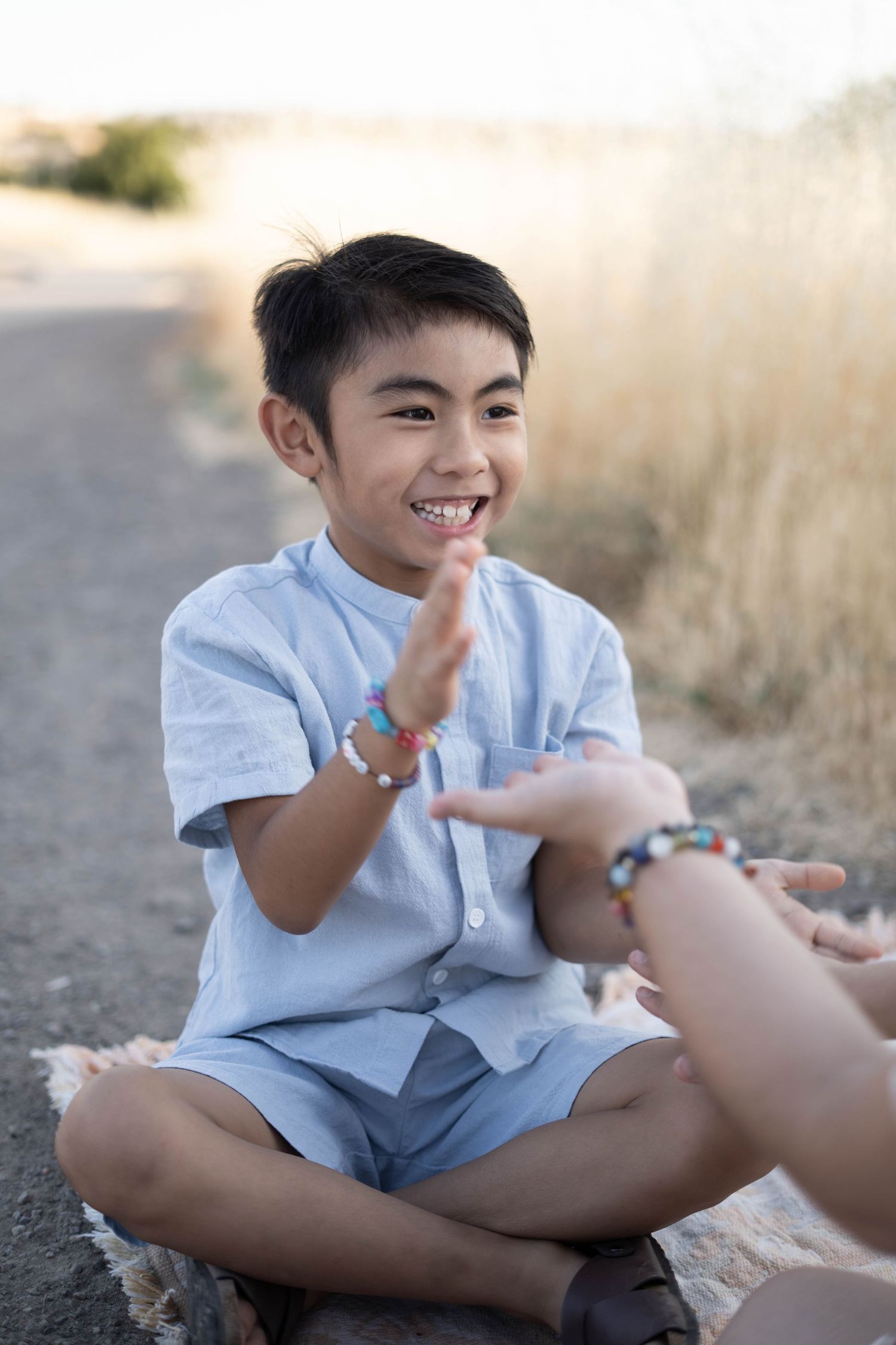 A child in light blue clothing sits cross-legged outdoors playing a hand game.